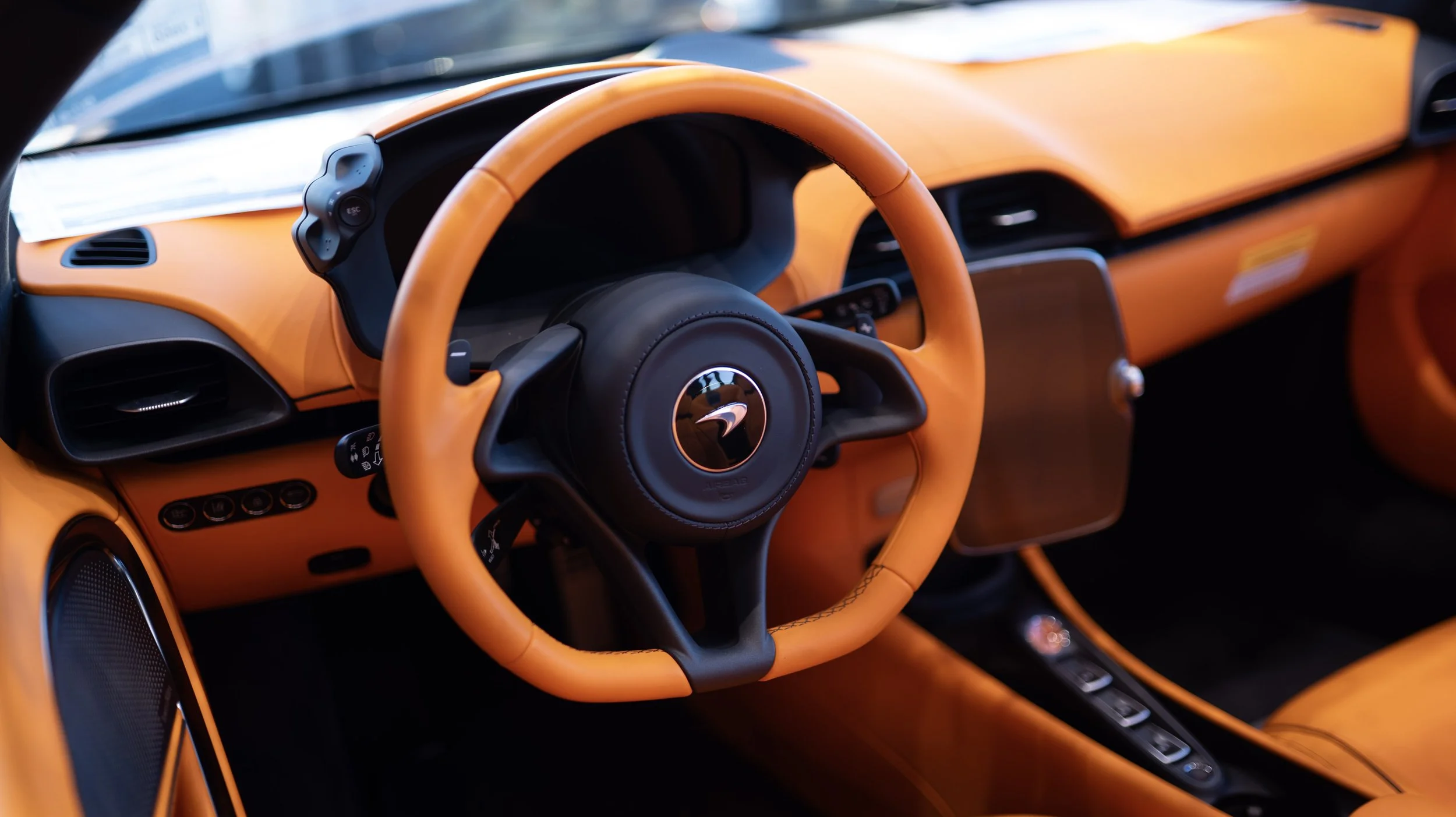 Interior view of a luxury sports car with bright orange interior, black steering wheel with logo, digital display screen, and control buttons.