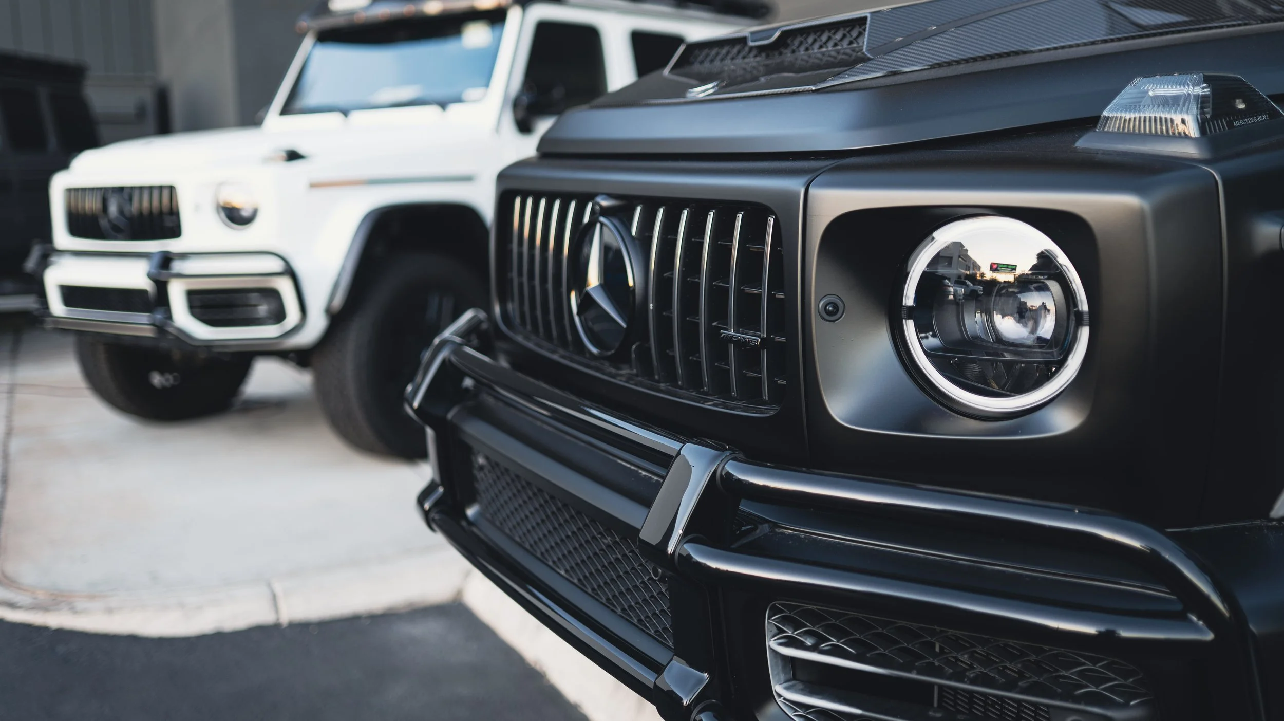 Close-up of the front grille and headlight of a black Mercedes-Benz G-Class SUV, with a white G-Class SUV blurred in the background.