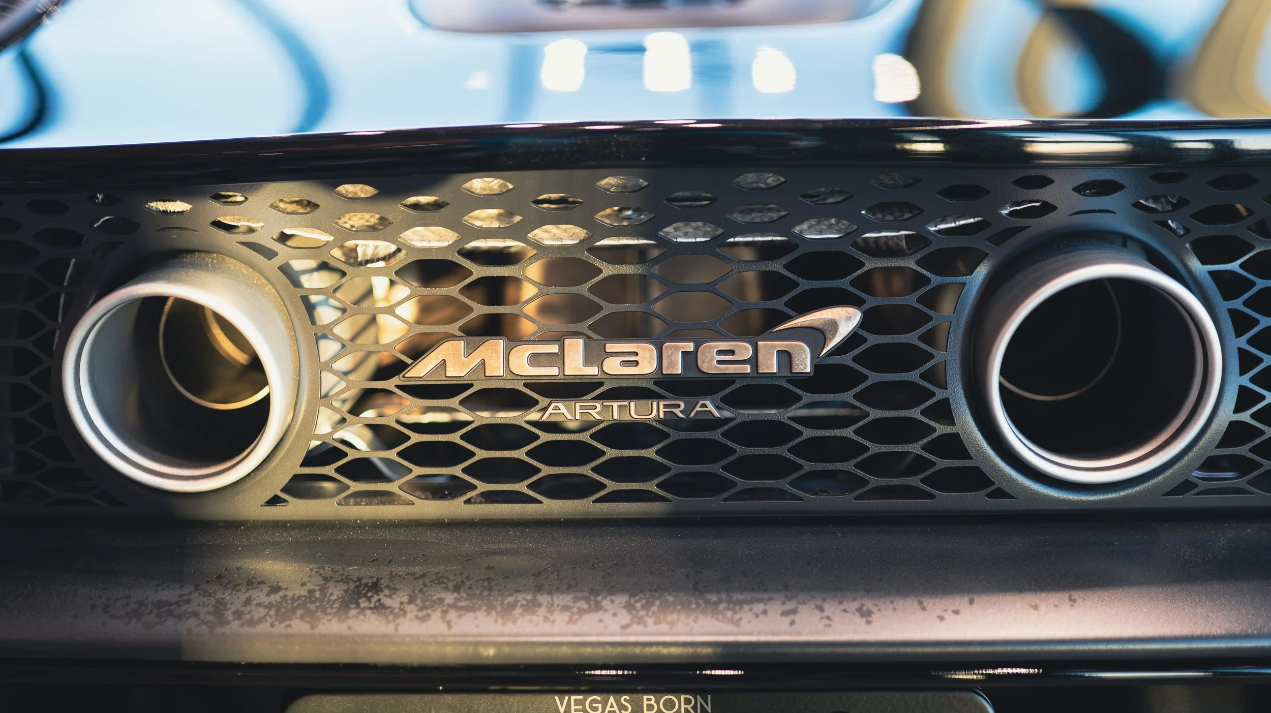 Close-up of a McLaren Artura emblem on the front grille of a sports car, with honeycomb pattern and dual circular air vents.