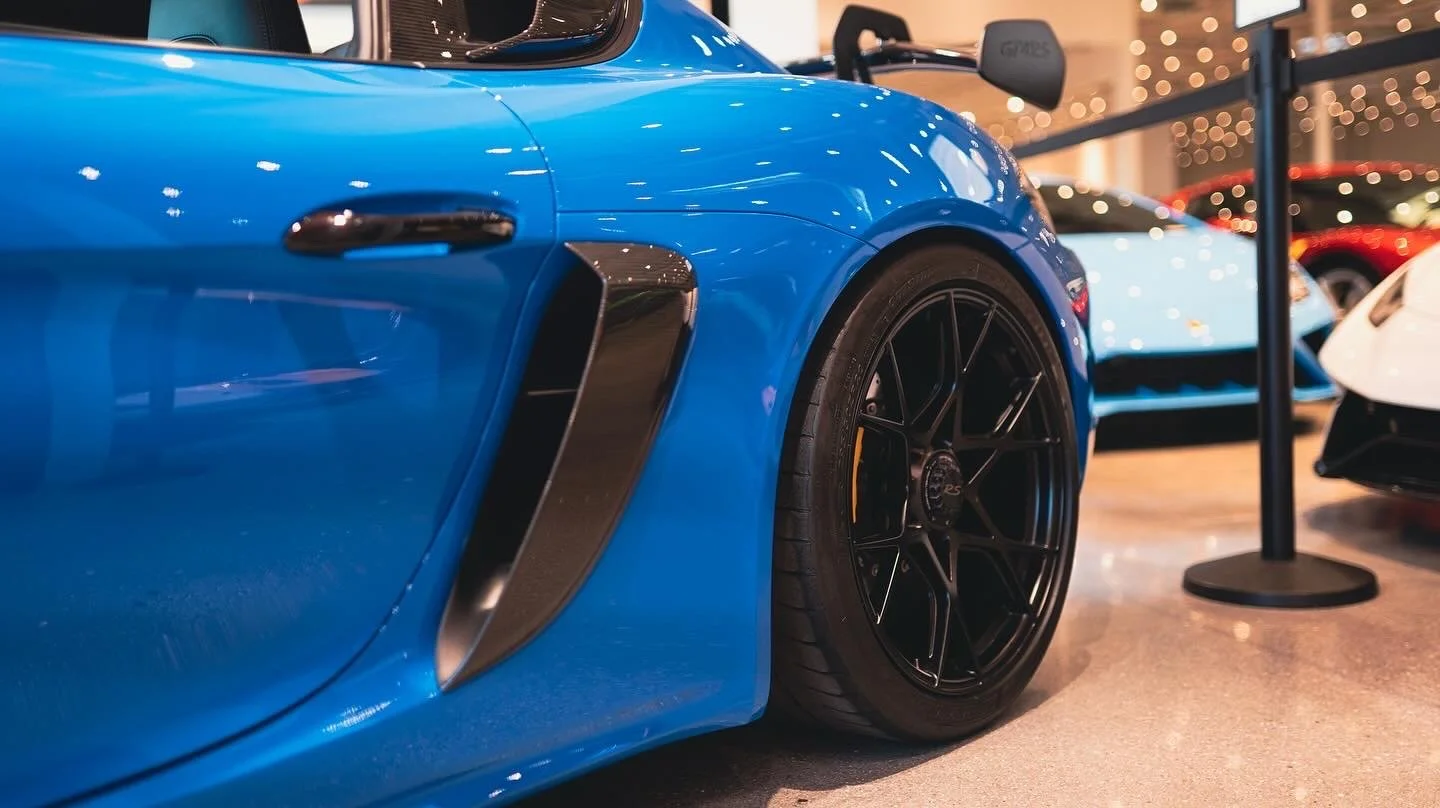 Close-up of a blue sports car at an indoor show, showing the front wheel, side vent, and part of the door, with other cars and decorative lights in the background.