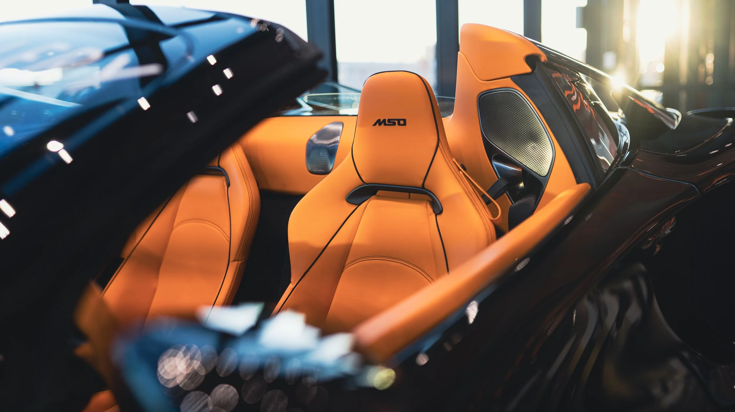 The interior of a luxury sports car featuring bright orange racing-style seats with black piping, a high-tech dashboard, and sunlight streaming through large windows.