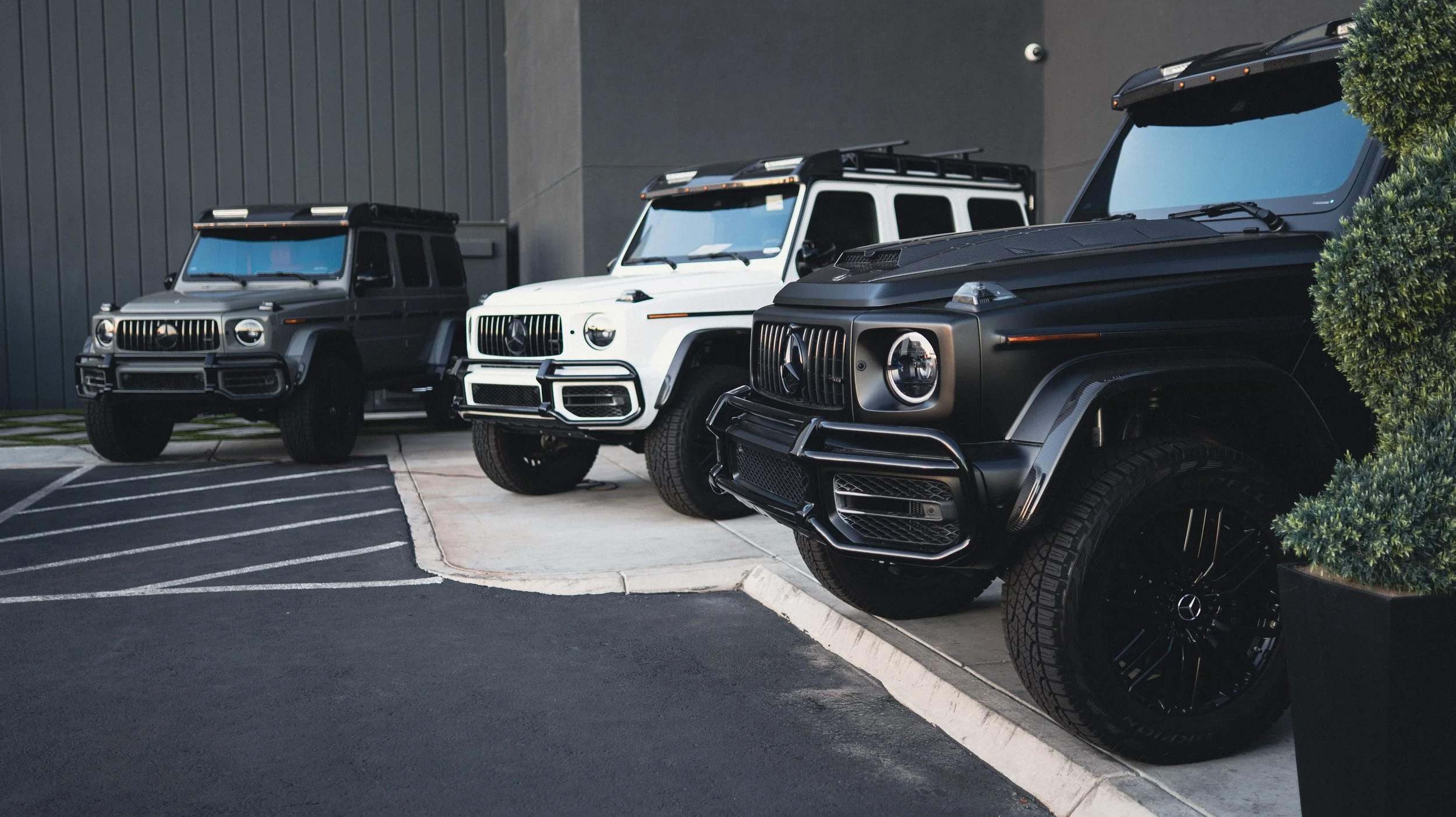 Three Mercedes-Benz G-Class SUVs parked outside near a gray building, with the black vehicle in the foreground, a white one in the middle, and another black vehicle farther back. There are manicured bushes on the right side. The parking lot surface is a mix of asphalt and concrete.