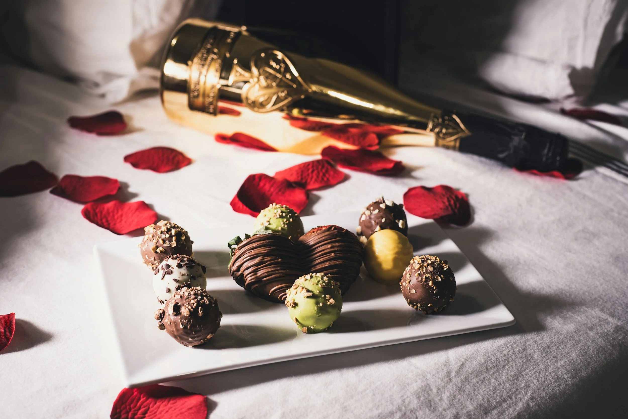 Photo of a hotel bed with red rose petals and a plate of chocolate covered strawberries and a bottle of champagne