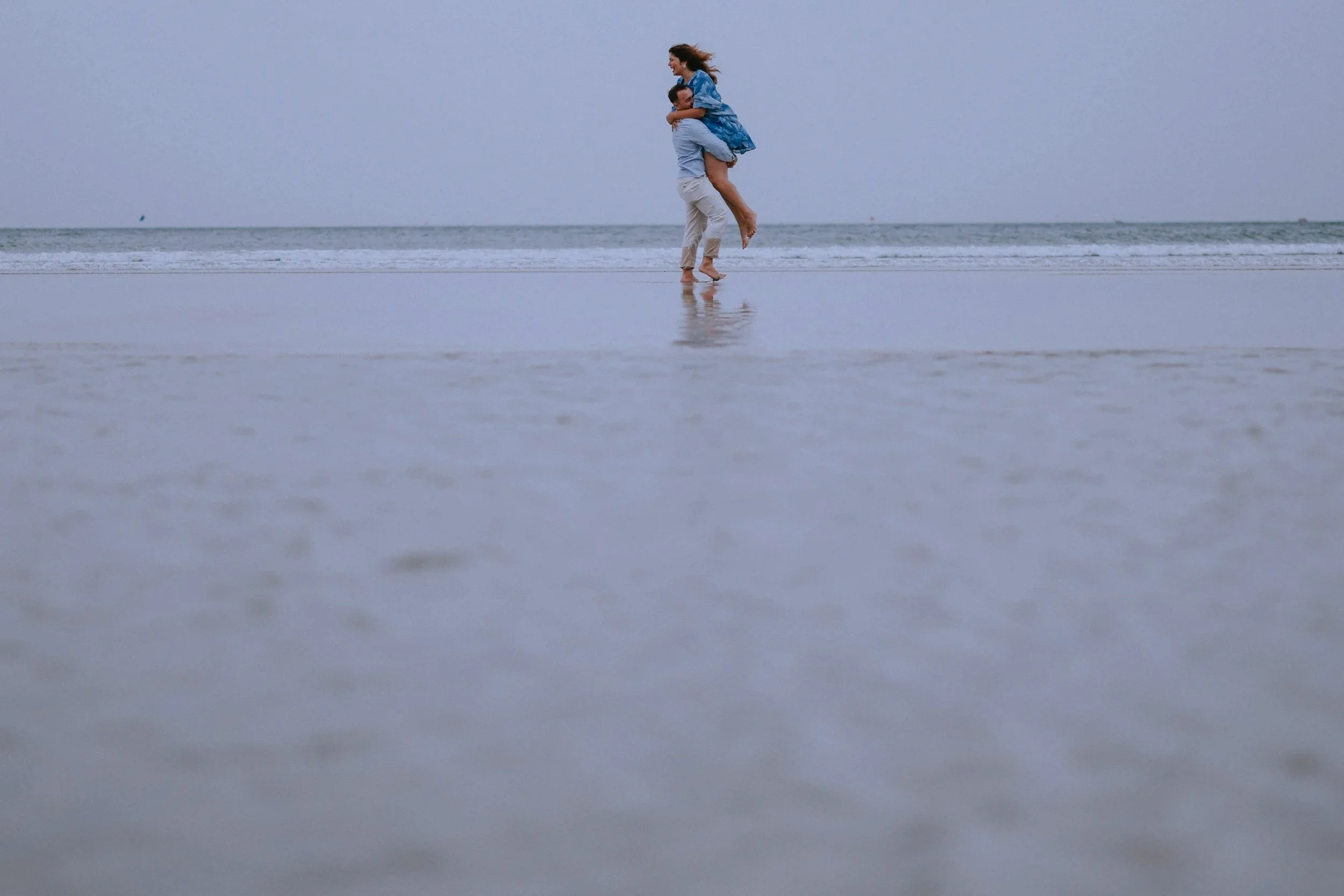 Couple on a beach, man lifting the woman up. The beach is empty and the sky is gray.