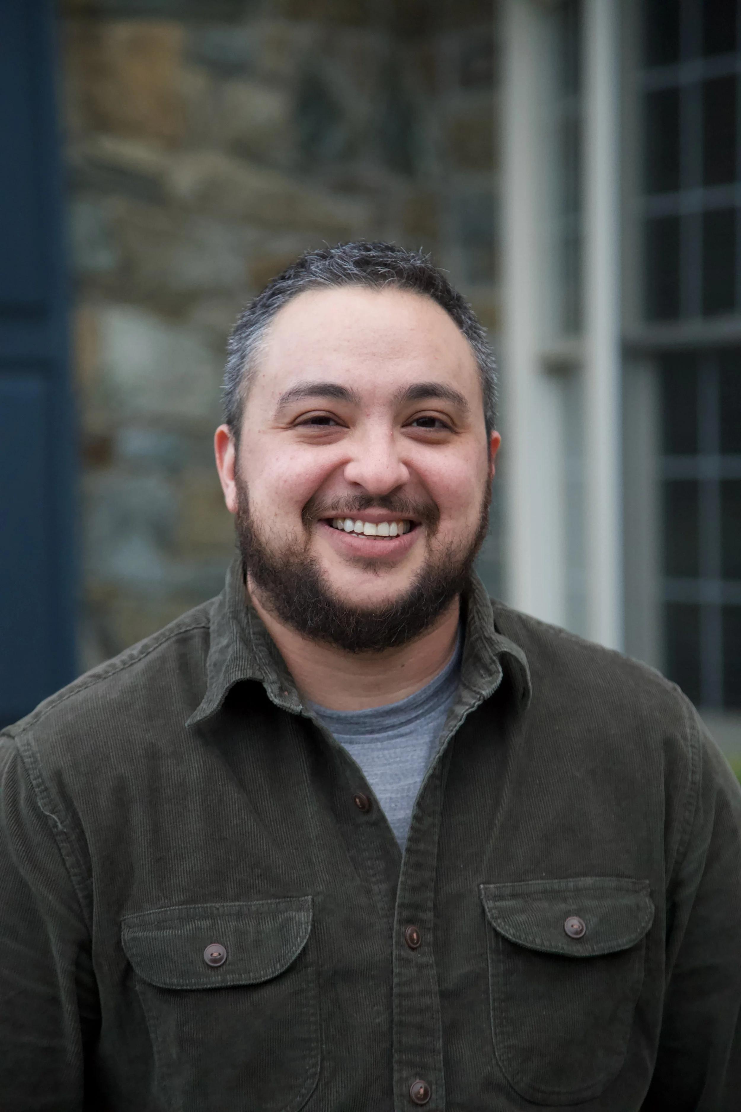 A smiling man with dark hair, a beard, and a mustache wearing a dark green button-up shirt over a gray T-shirt, standing outdoors with a stone building and windows in the background.