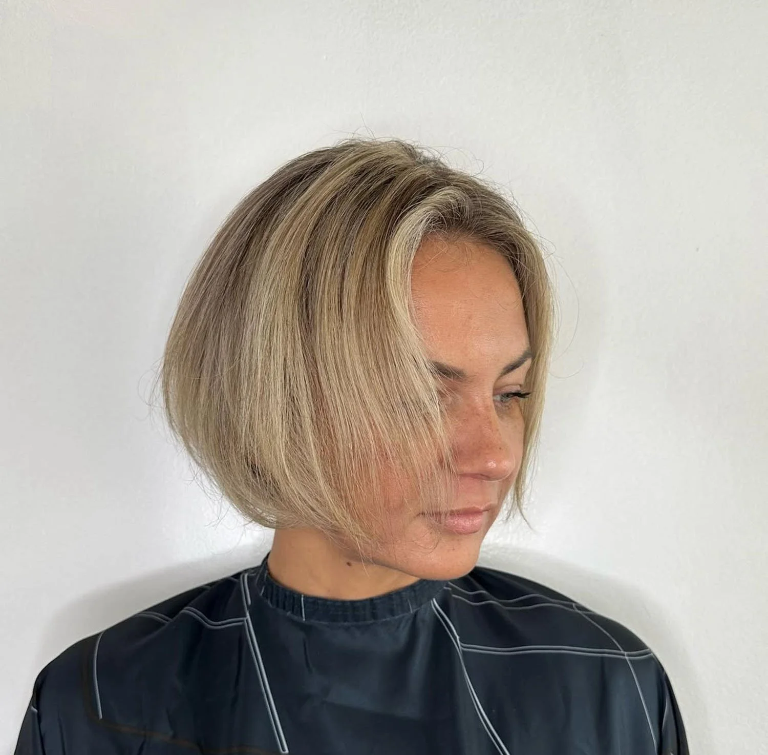 Woman with chin-length blonde hair, styled in a sleek, layered bob, wearing a black salon cape with white stitching, standing against a plain white wall.