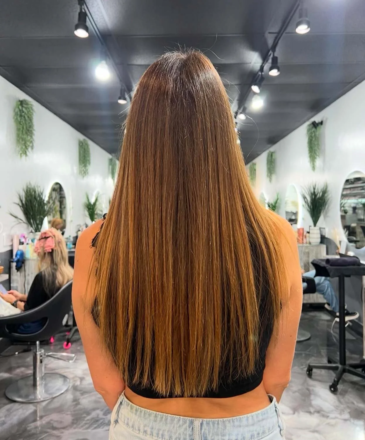 Woman with long straight light brown hair in a salon, with mirrors and plants on the white walls.