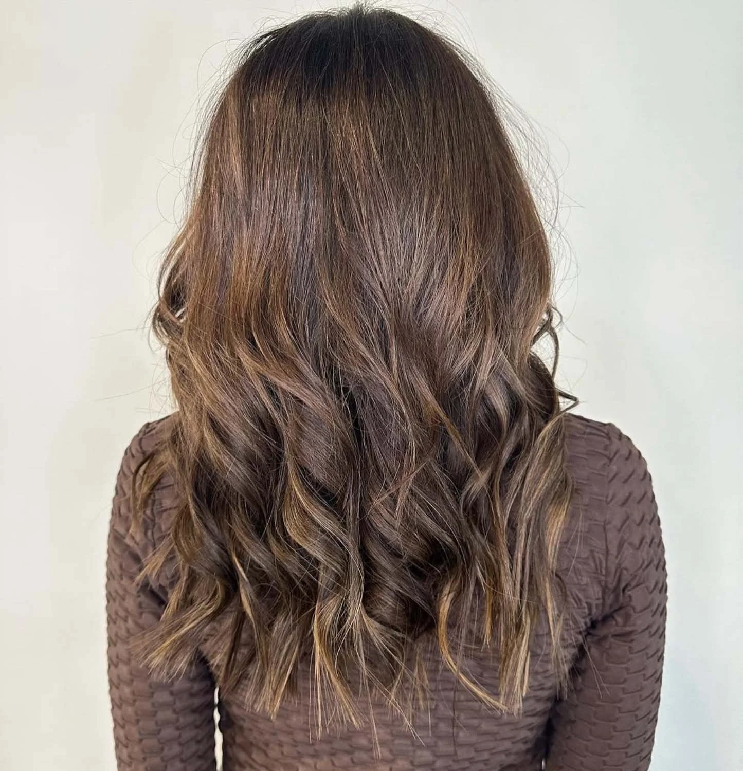Back view of a woman with long, wavy brown hair wearing a textured brown top, standing against a plain white background.