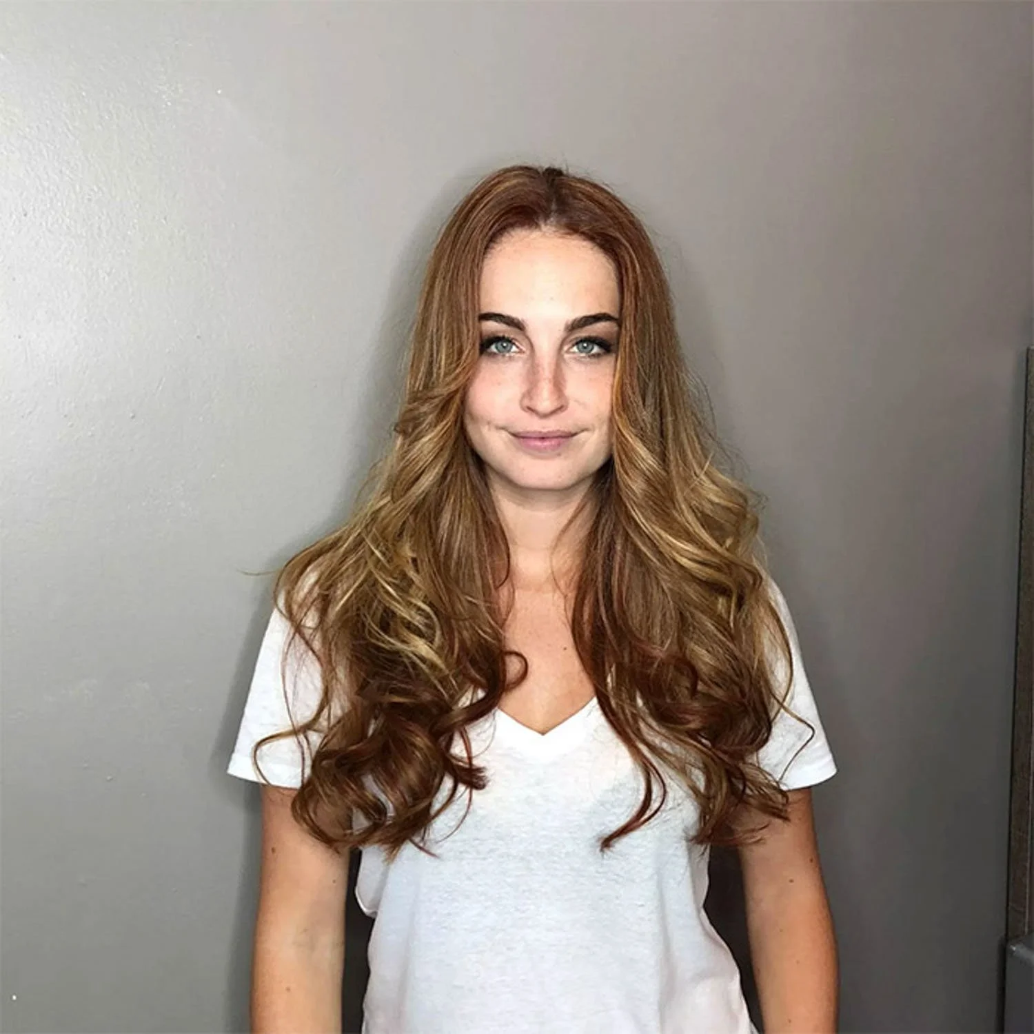 A woman with long, wavy red hair wearing a white V-neck T-shirt standing against a plain gray wall.