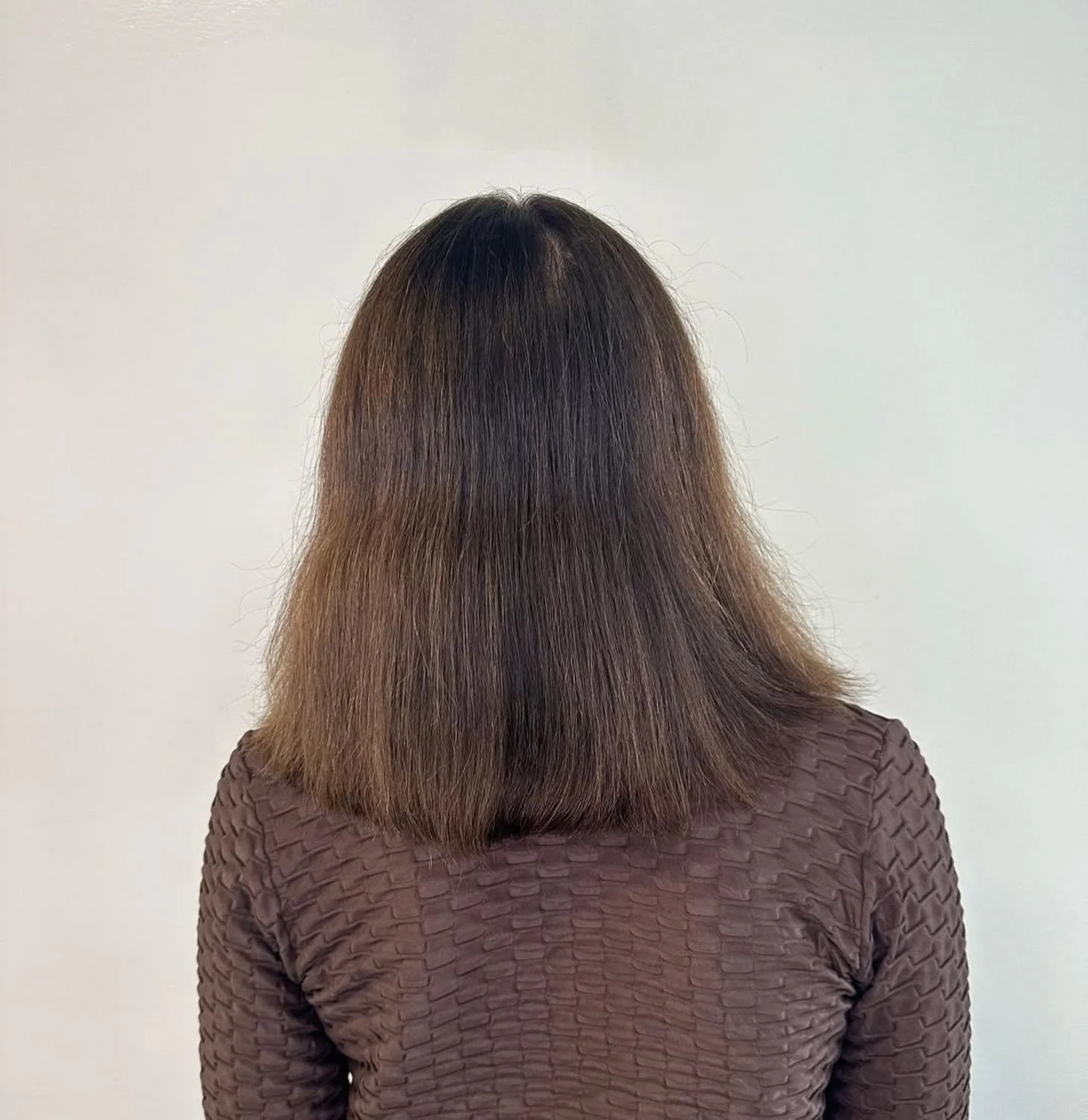 Back view of a woman with medium-length brown hair wearing a textured brown top, standing against a plain white background.