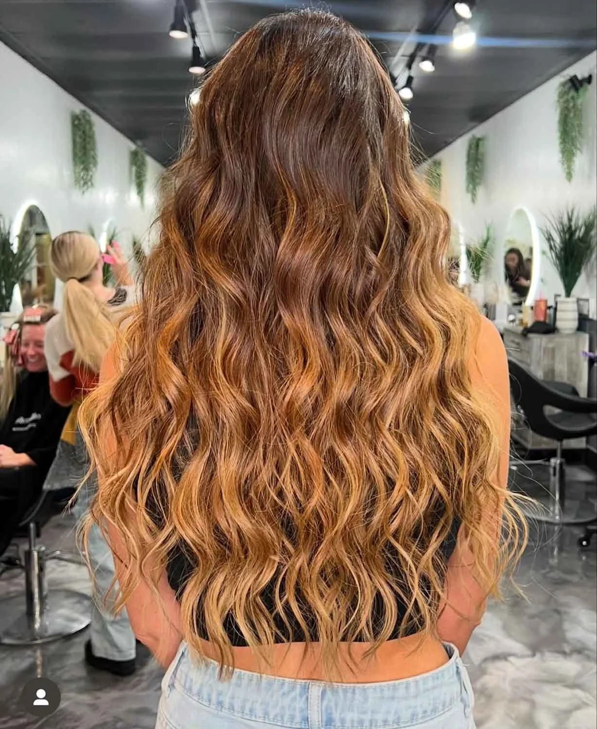 Back view of a woman with long, curly, ombre hair in shades of brown and blonde, inside a hair salon with mirrors, plants, and people in the background.