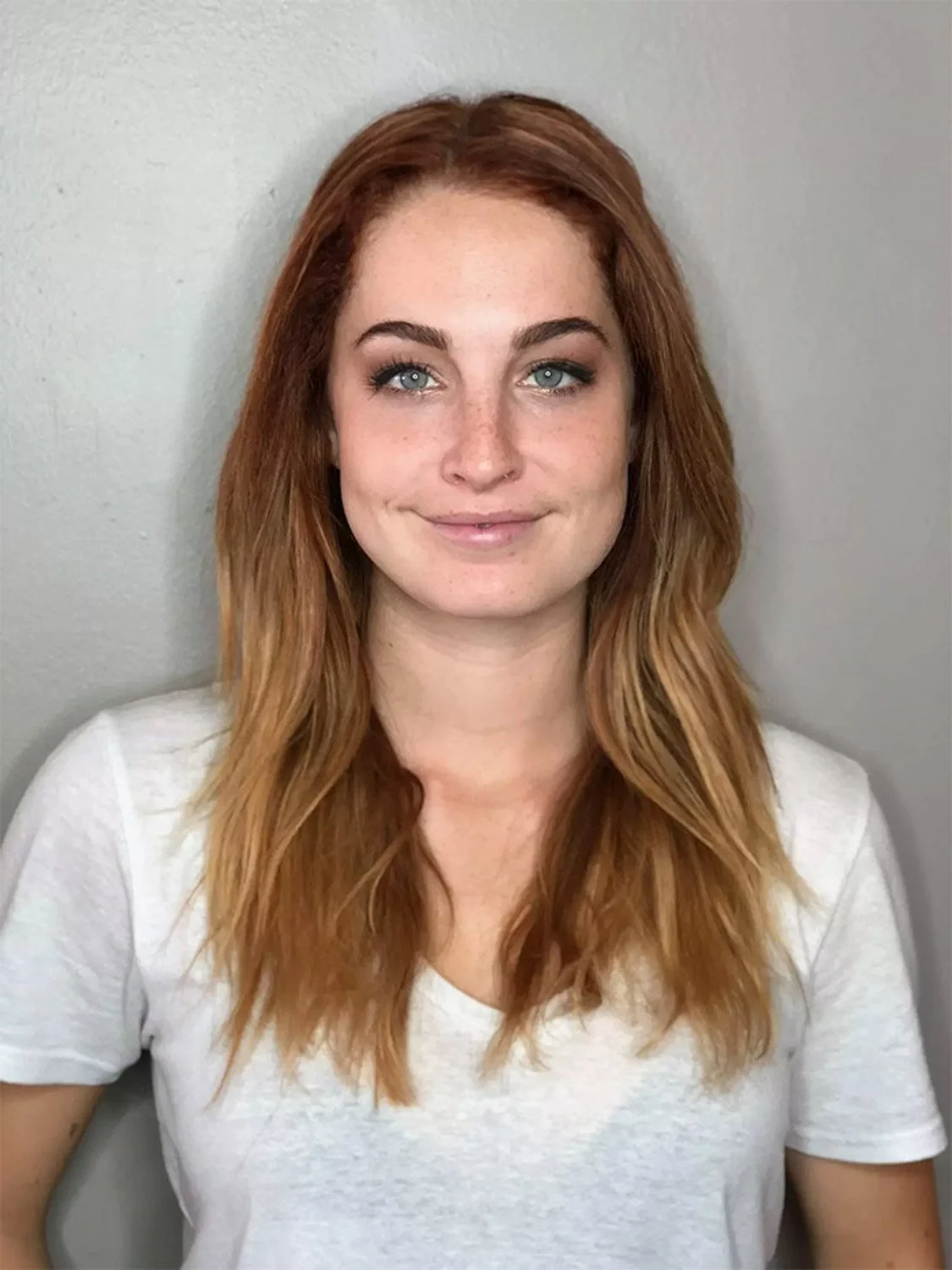 A young woman with long red hair, blue eyes, and fair skin, smiling softly at the camera, wearing a white T-shirt, against a plain gray background.