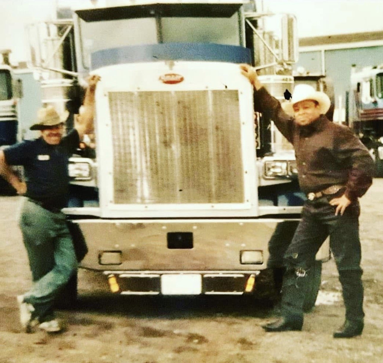 Two men wearing cowboy hats posing next to a large commercial truck, with one man leaning on the truck's front grille and the other standing with his hand on his hip, in an outdoor lot with other trucks in the background.