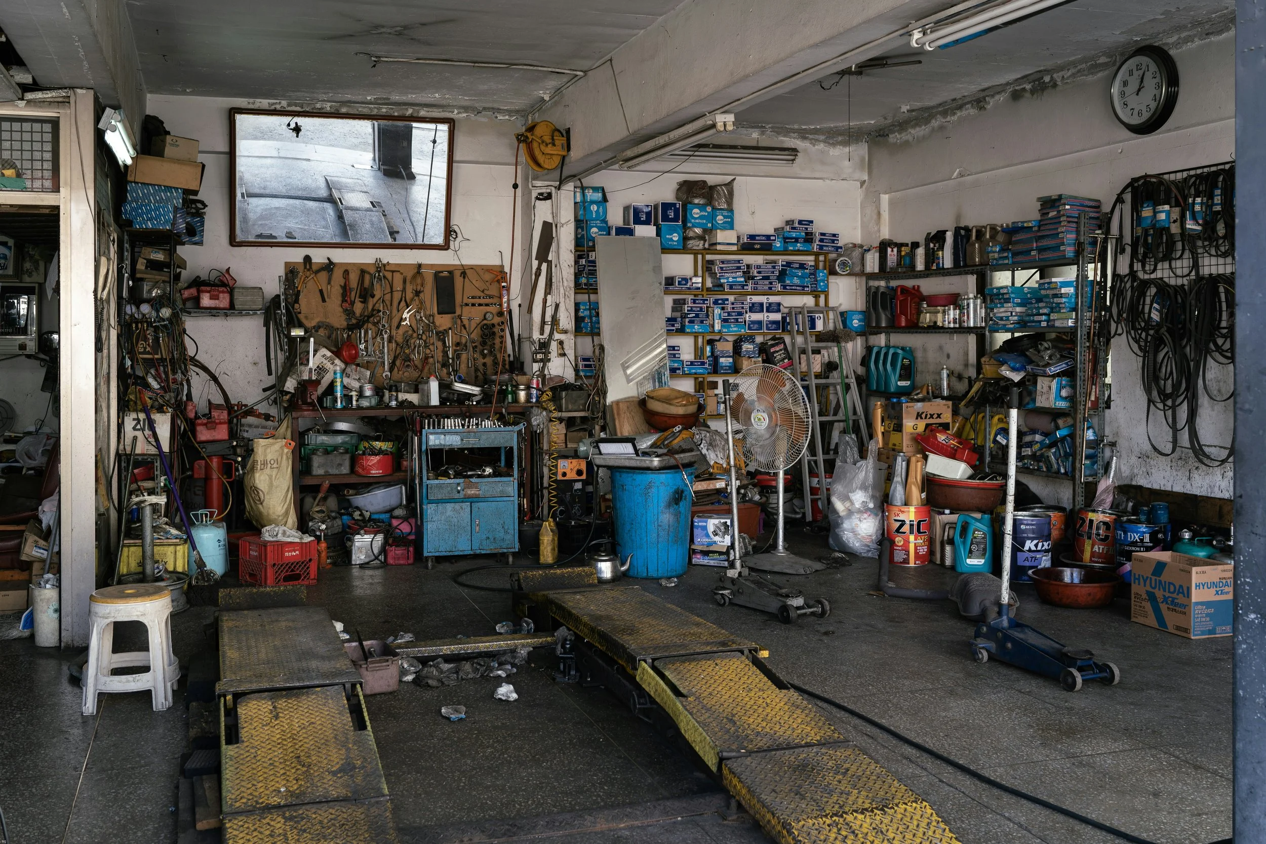 A cluttered auto repair garage with tools, shelves, and repair equipment, including car lift ramps, workbenches, spare parts, and tools.
