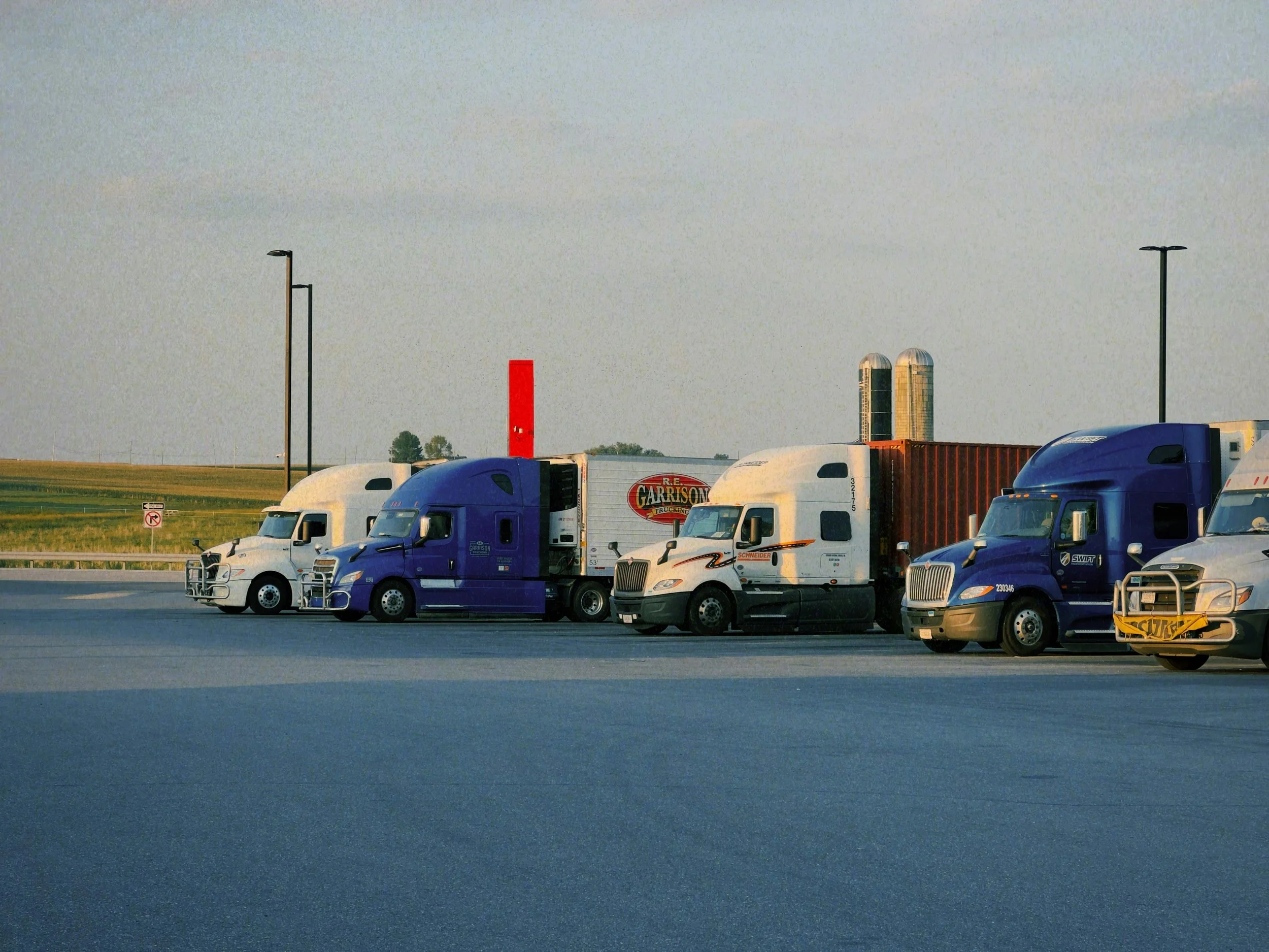 Line of semi-trucks parked on a highway with fields in the background.