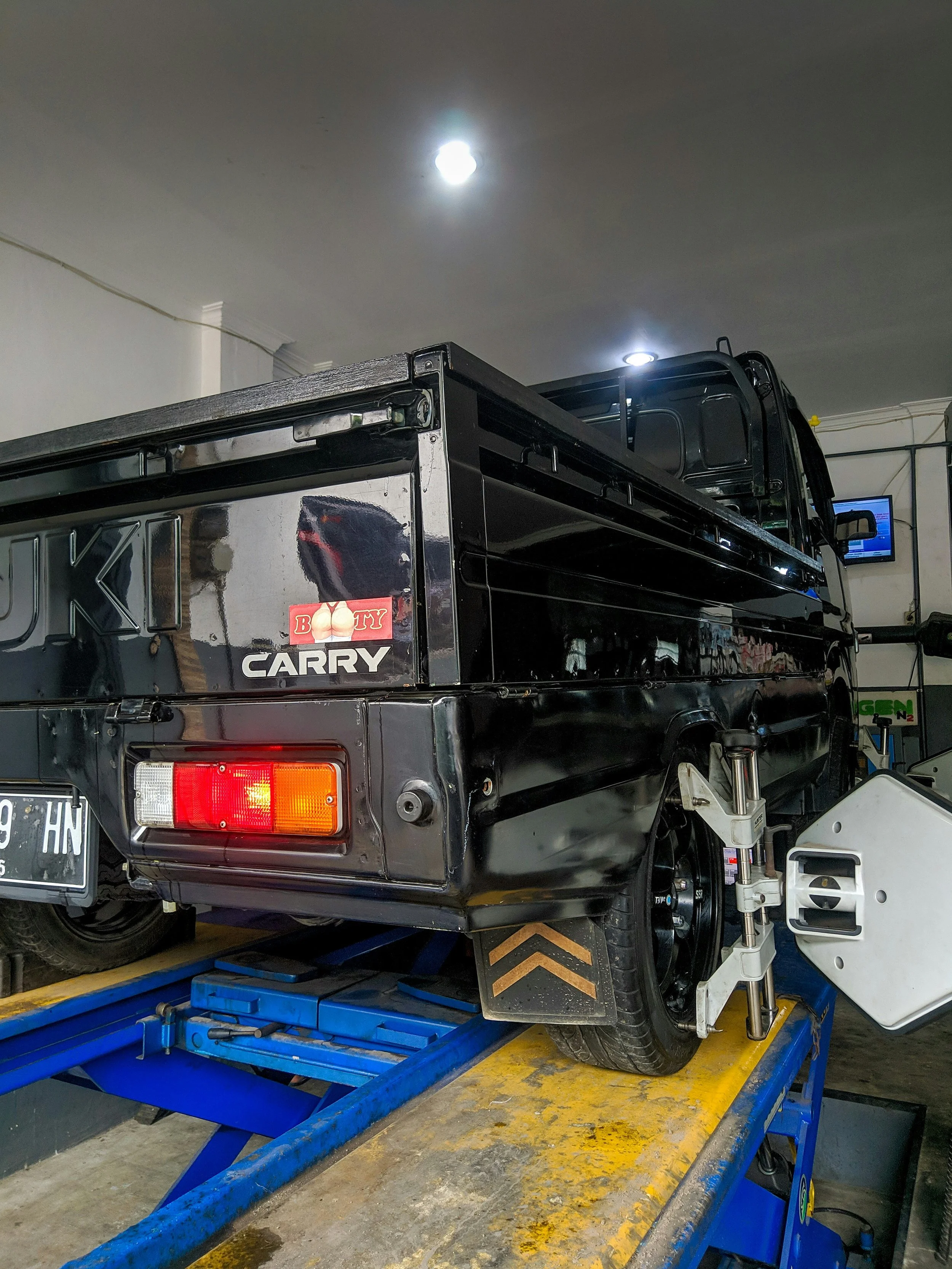 A black pickup truck with a sticker that says 'Booty' and an image of a person's buttocks, parked on a yellow and blue platform inside a garage.