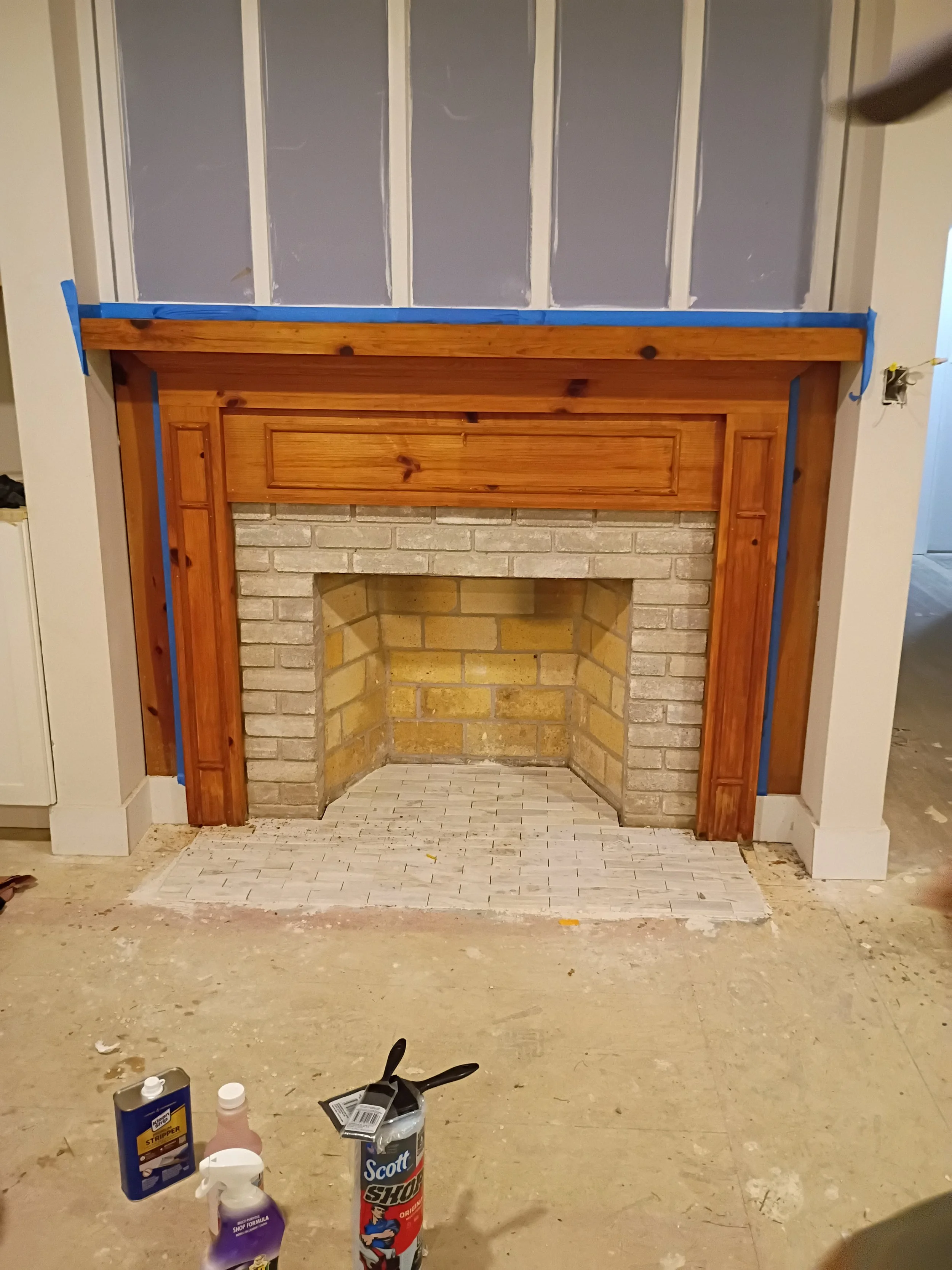 A brick fireplace with a wooden mantel in a room under renovation, with construction supplies on the floor.