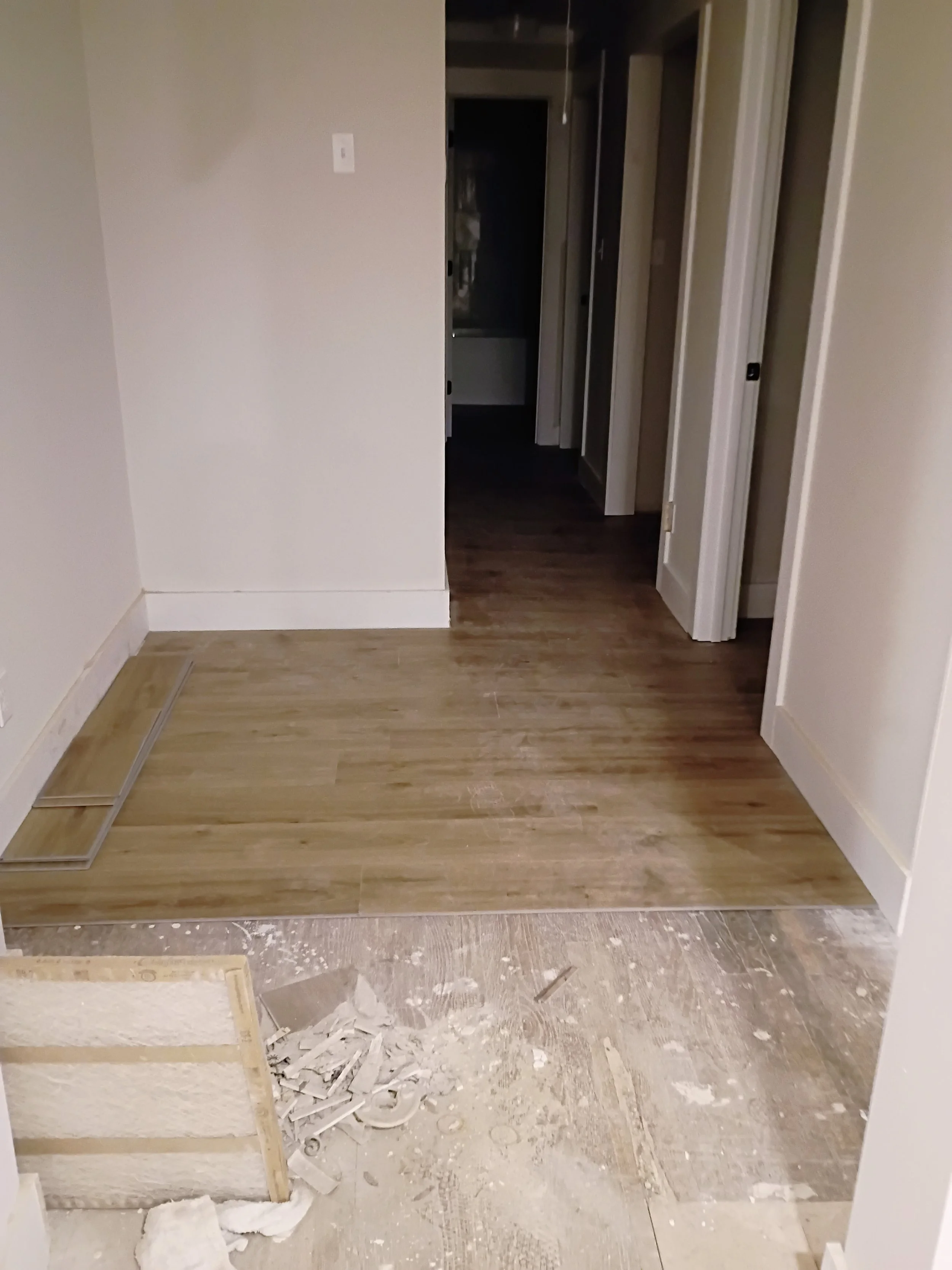 Room with partially installed hardwood flooring and construction debris in the foreground, hallway with closed doors leading to other rooms.