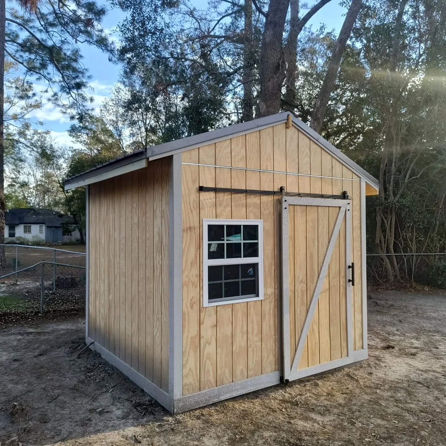 A small wooden shed with a single window and a sliding barn door, situated on a dirt ground with trees and a chain-link fence in the background.