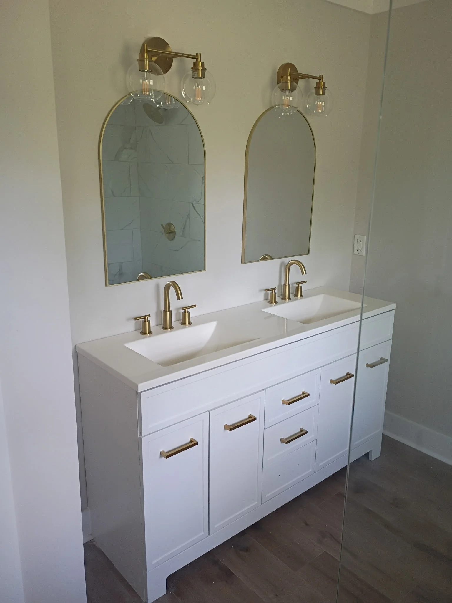 White double vanity with gold handles and fixtures, two oval mirrors, and modern lighting with exposed bulbs, in a contemporary bathroom.