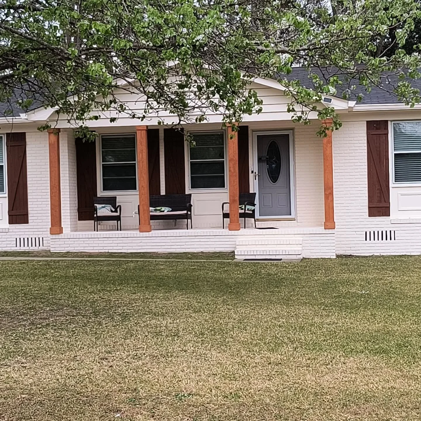 Front porch of a house with a brick foundation, four wooden pillars, two chairs, a bench, and a gray front door, with windows and shutters, and a tree in the top part of the image.