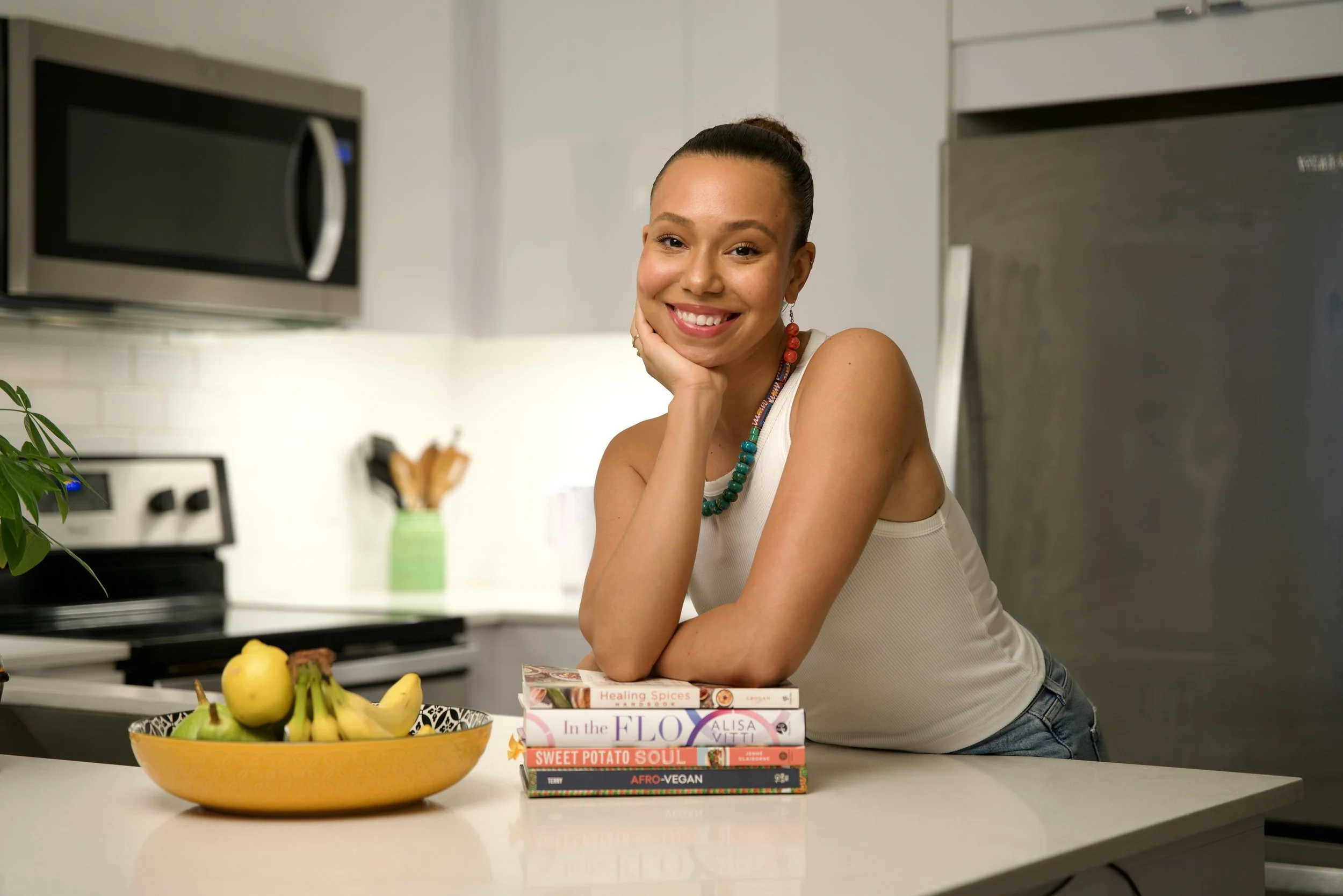 A young woman smiles and rests her chin on her hand in a modern kitchen, with a bowl of bananas and a stack of books on the counter.