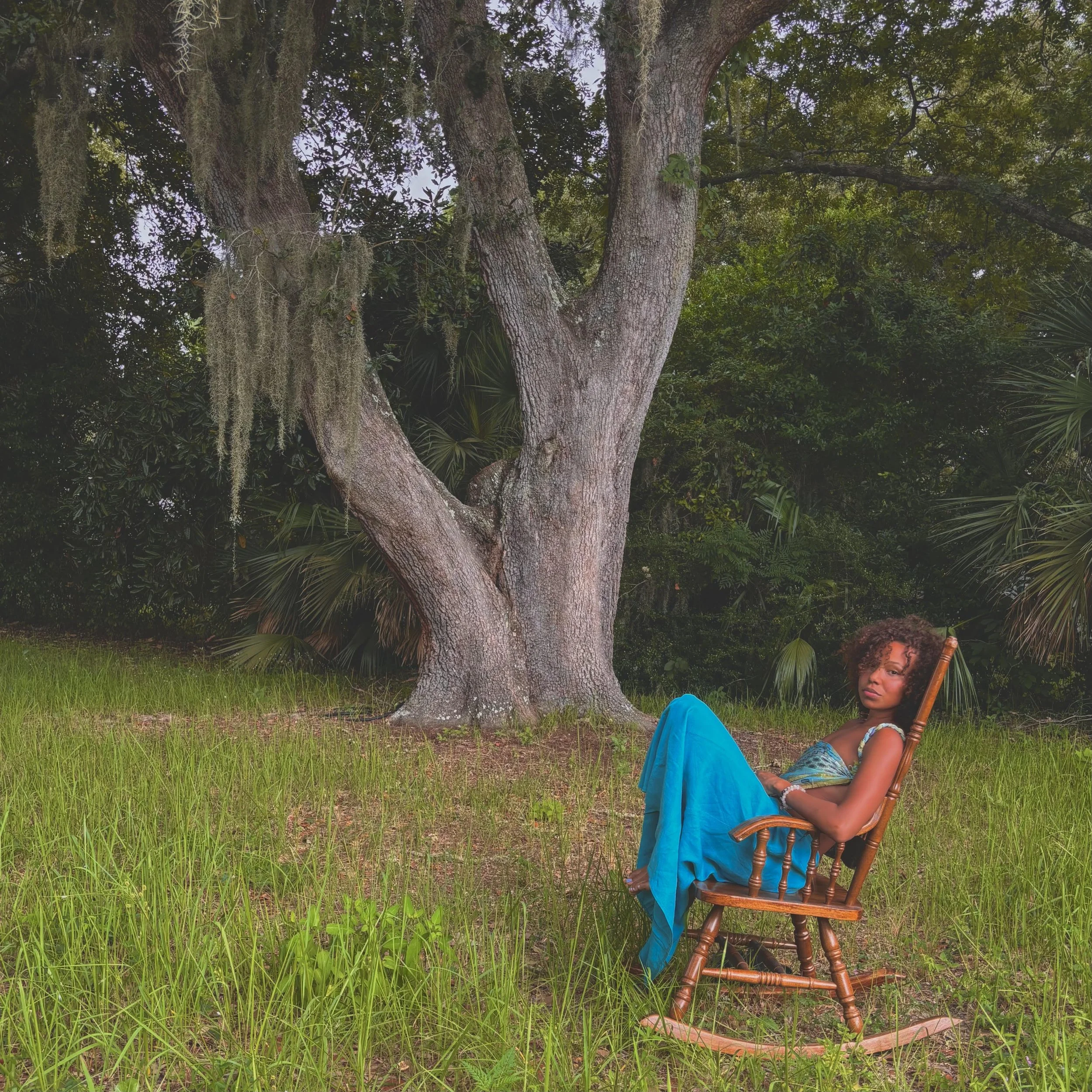 A woman sitting in a wooden rocking chair outdoors under a large tree with hanging moss and dense foliage in the background, wearing a blue dress.