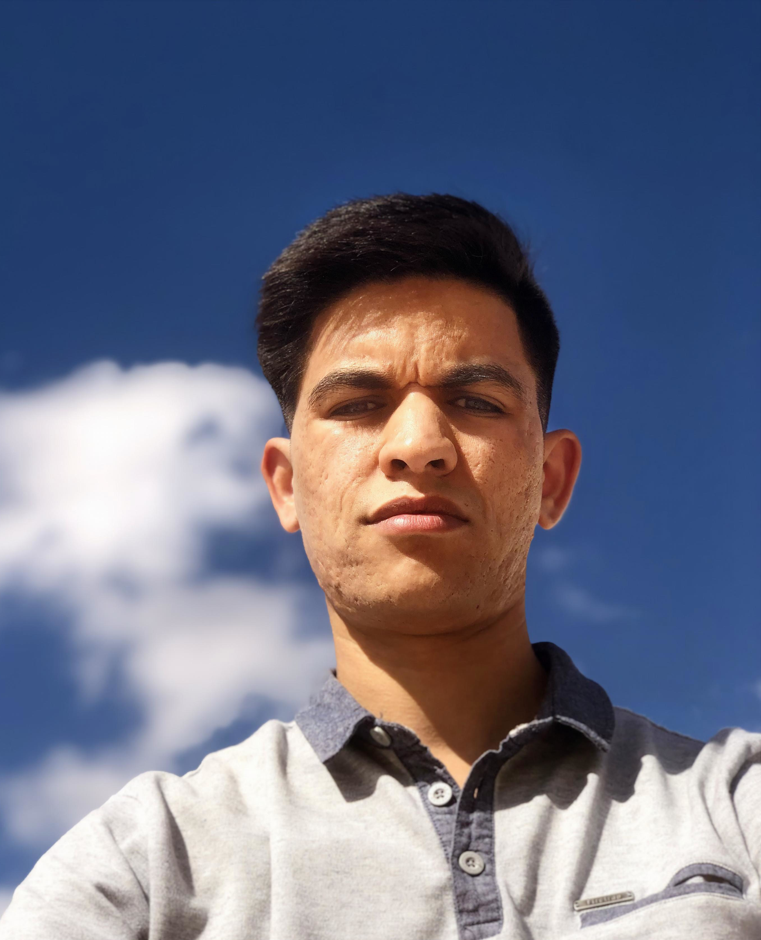 A young man taking a selfie outdoors against a blue sky with a few clouds.
