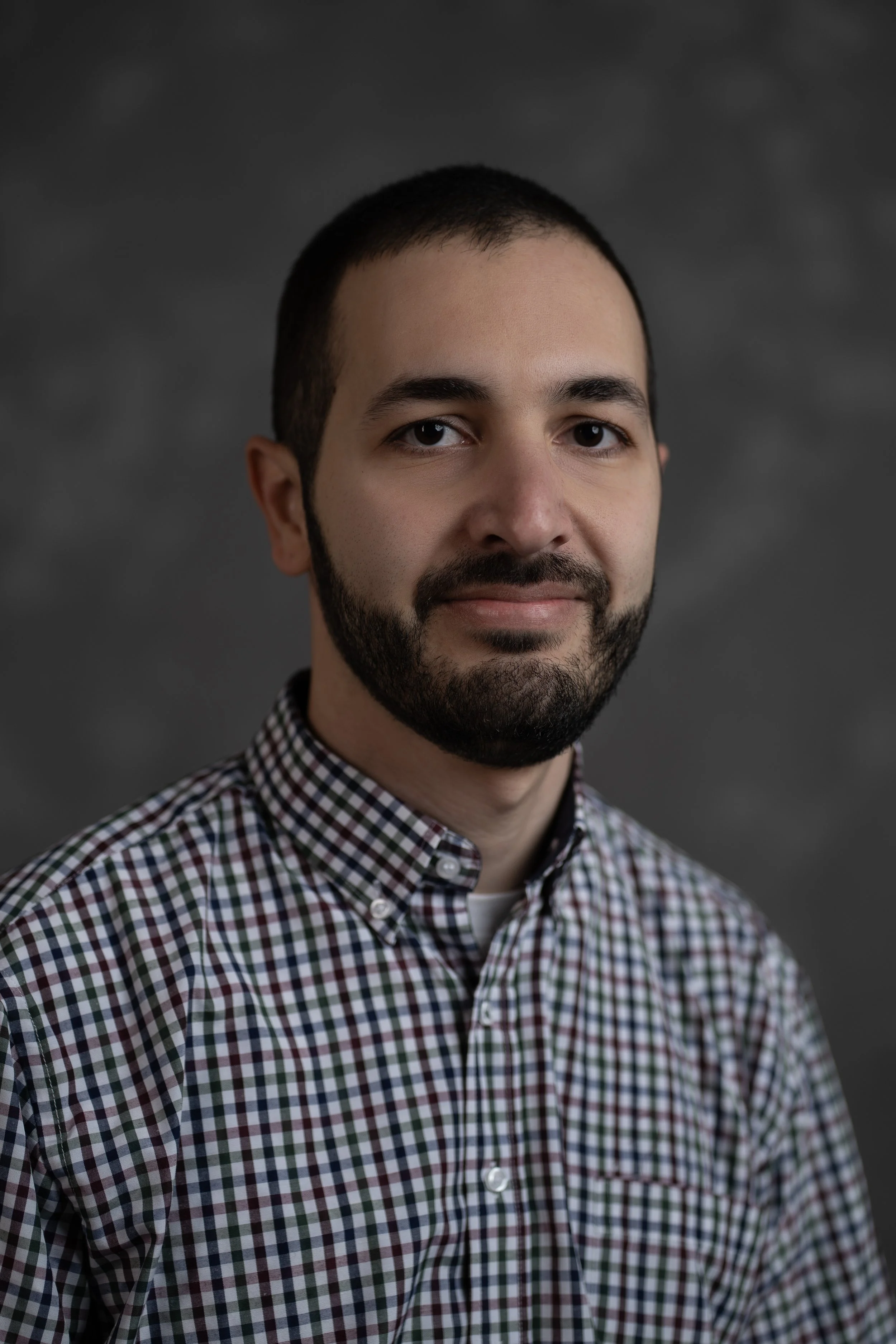Portrait of a man with a beard wearing a checkered shirt, against a dark gray background.