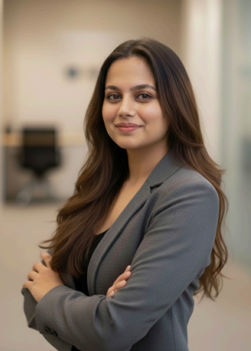 A woman with long brown hair, wearing a gray blazer over a black top, standing confidently with arms crossed in a professional indoor setting.