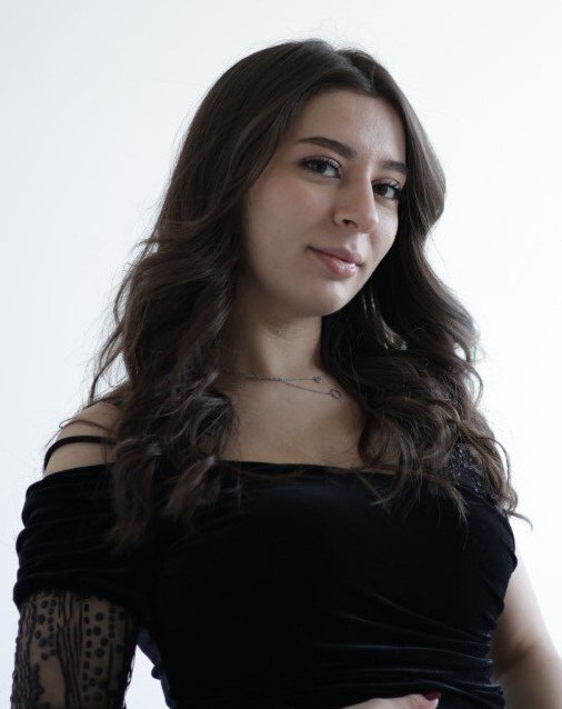 Young woman with long wavy brown hair wearing a black off-the-shoulder top standing against a white background.