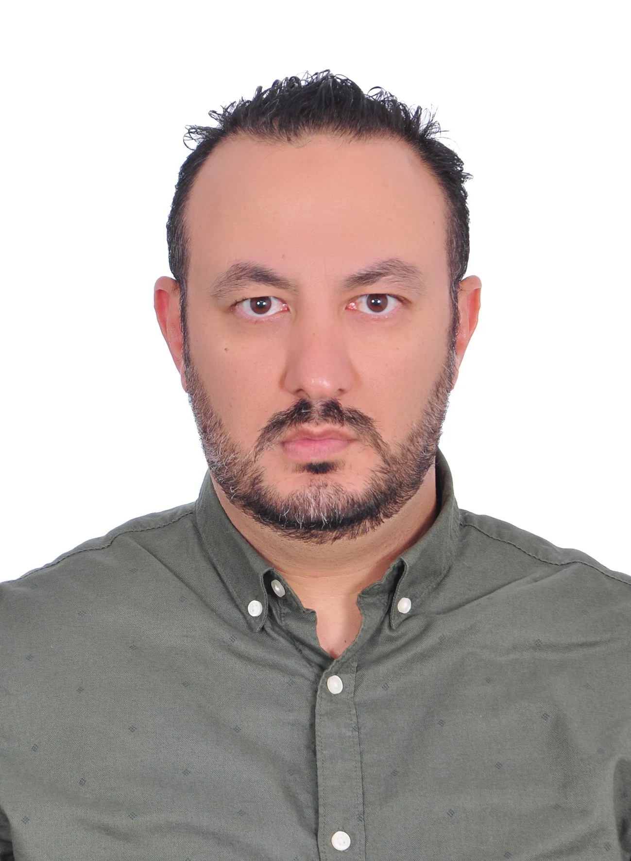 Close-up portrait of a man with dark hair, a beard, and wearing a green button-up shirt, looking directly at the camera with a serious expression, against a plain white background.