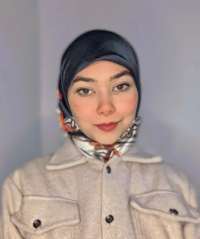 A young woman wearing a dark headscarf with a patterned scarf underneath, smiling at the camera. She is dressed in a light-colored jacket with visible buttons and pockets, standing against a plain background.