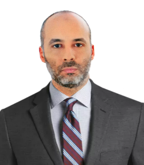 A man with a closely shaved head and beard, wearing a dark gray suit, light blue dress shirt, and a striped tie, looking directly at the camera against a plain white background.