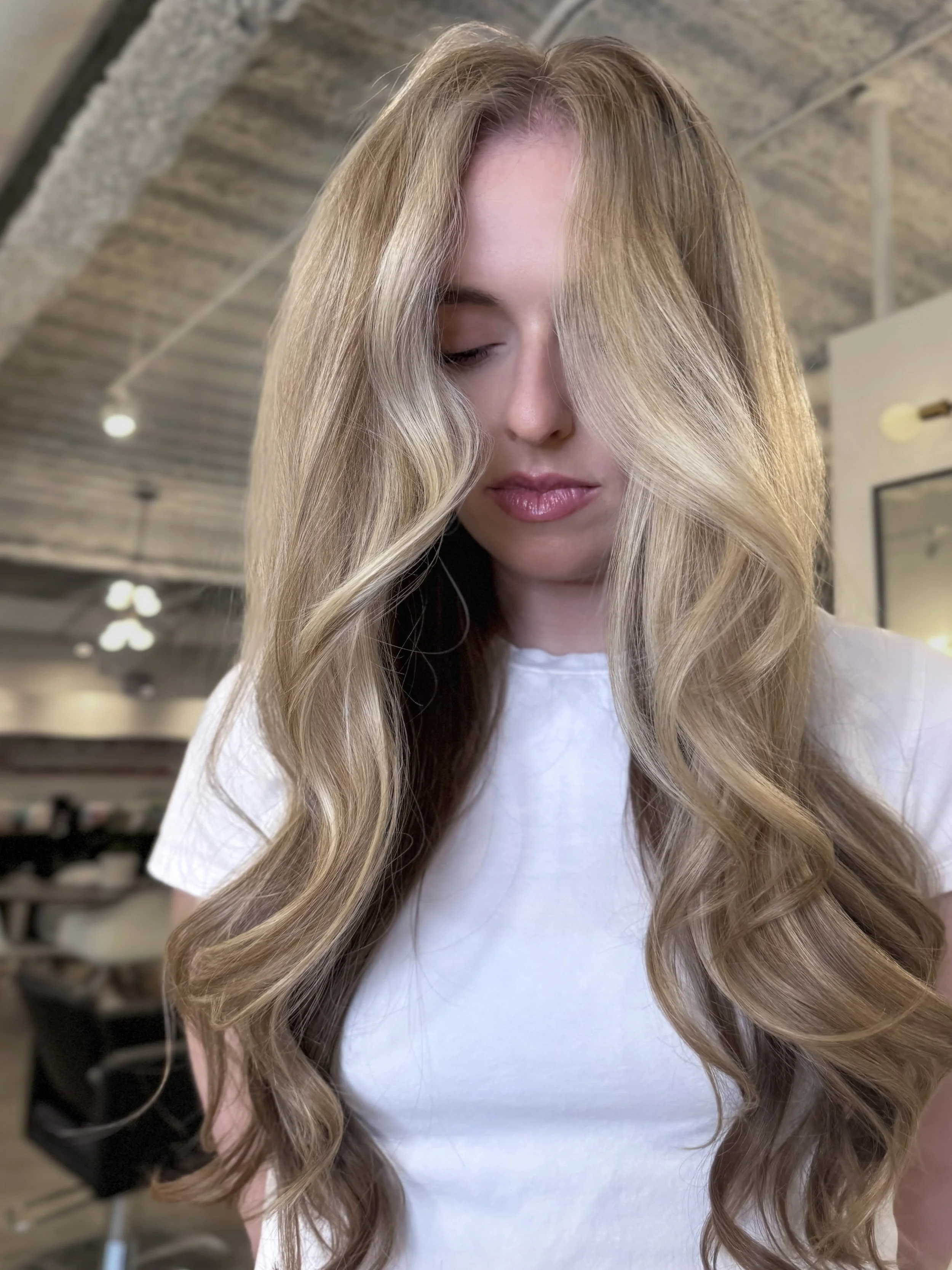 Close-up of a woman with long, wavy blonde hair and closed eyes, wearing a white top, in a public setting.