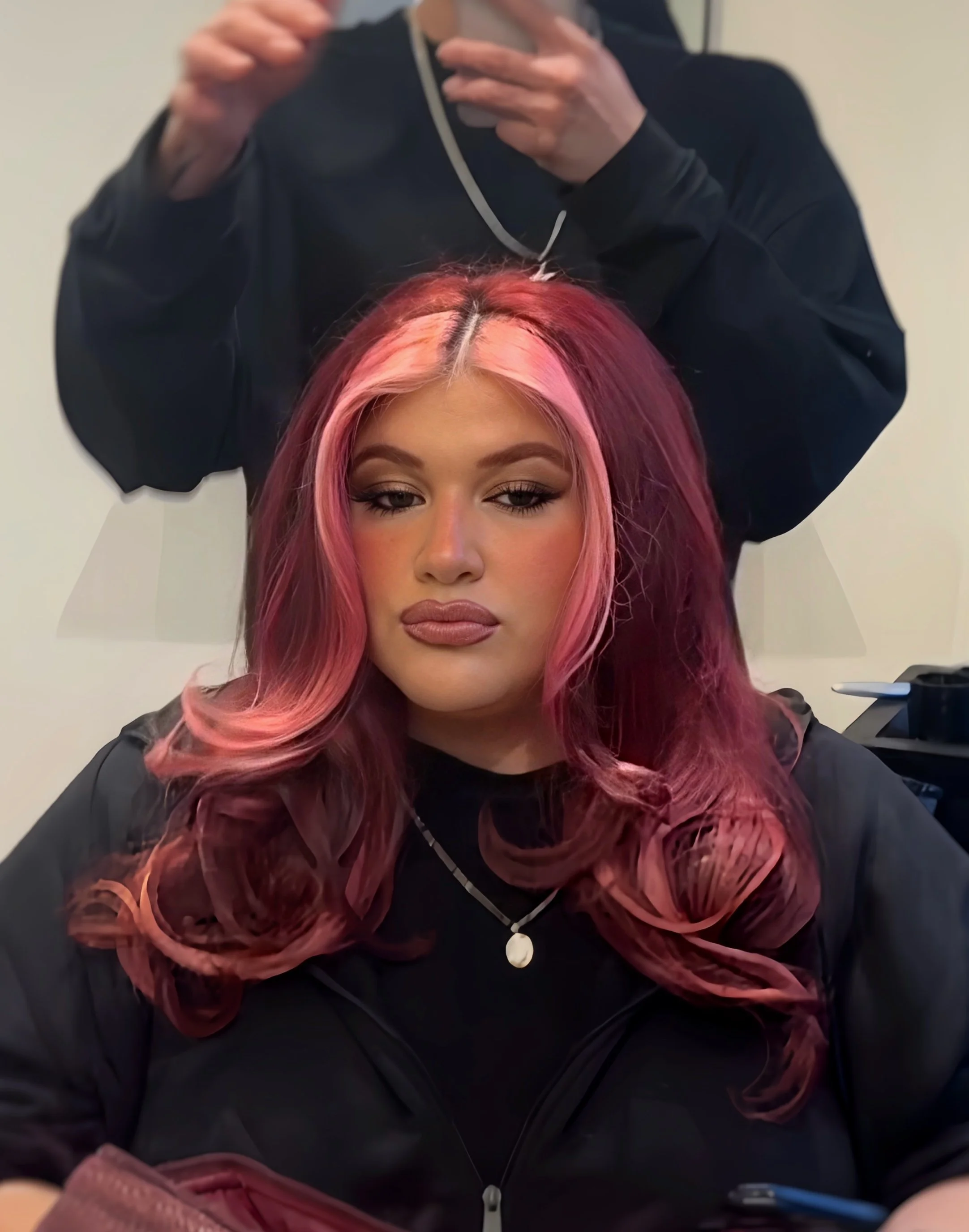 A woman with pink and purple hair, wearing makeup, sits in front of a mirror while a hairstylist with a black jacket and face mask adjusts her hair in a salon.