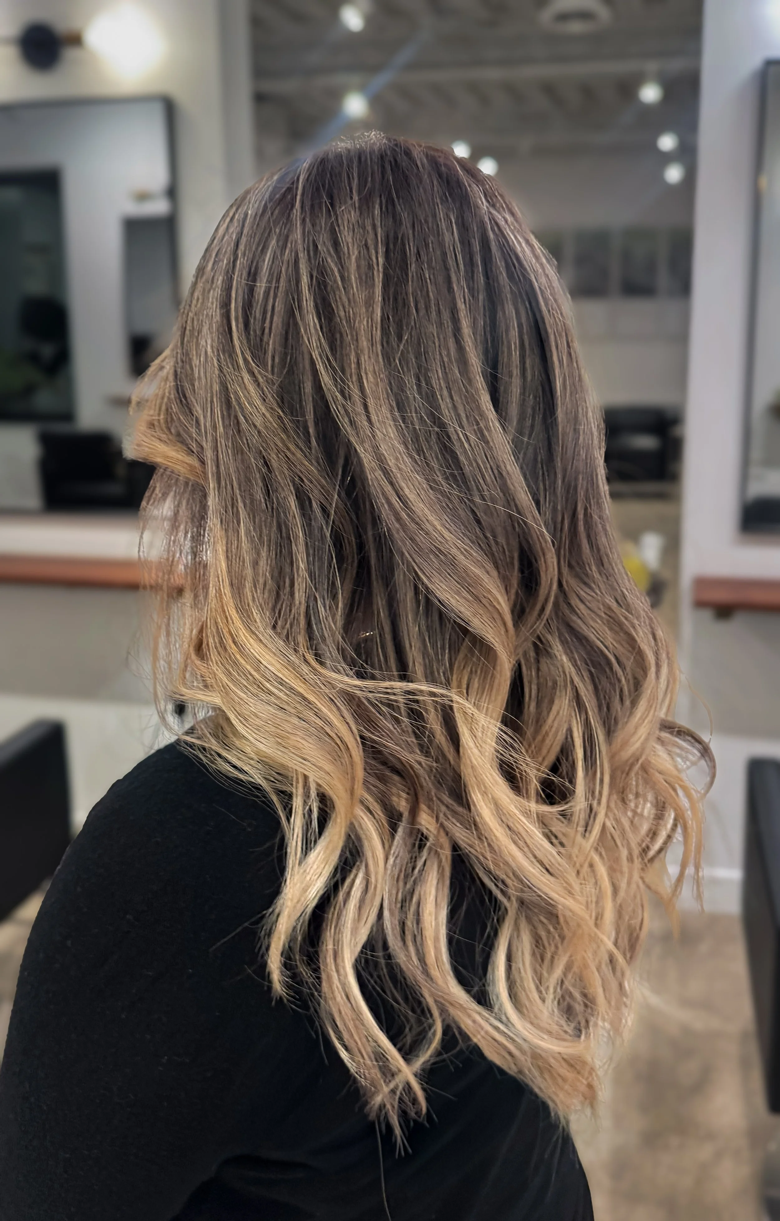 A woman with long, wavy hair that is highlighted in blonde shades, wearing a black top indoors with a modern interior setting.