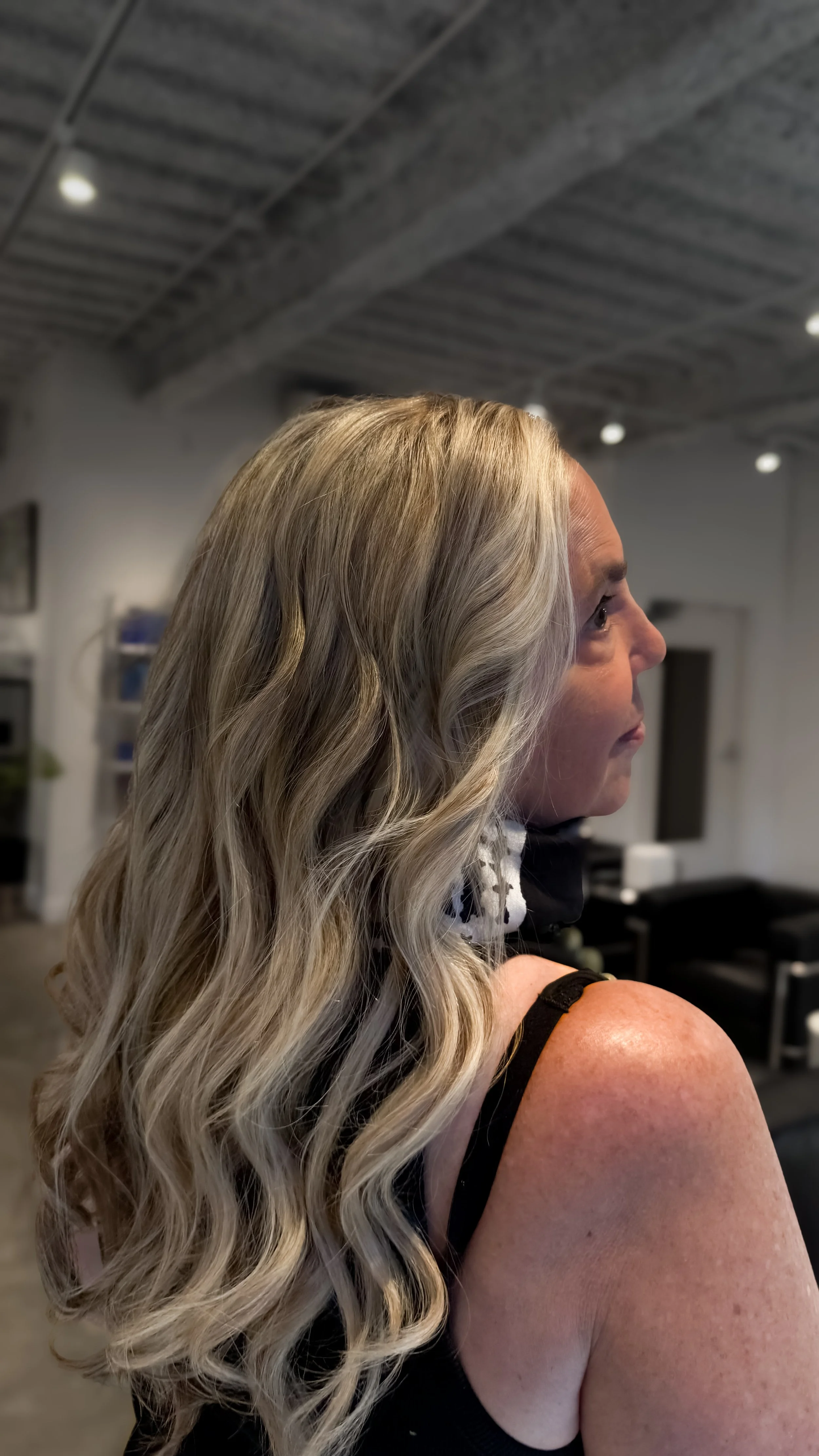 A woman with long, wavy blonde hair is seen in profile inside a salon with a gray ceiling and salon equipment in the background.