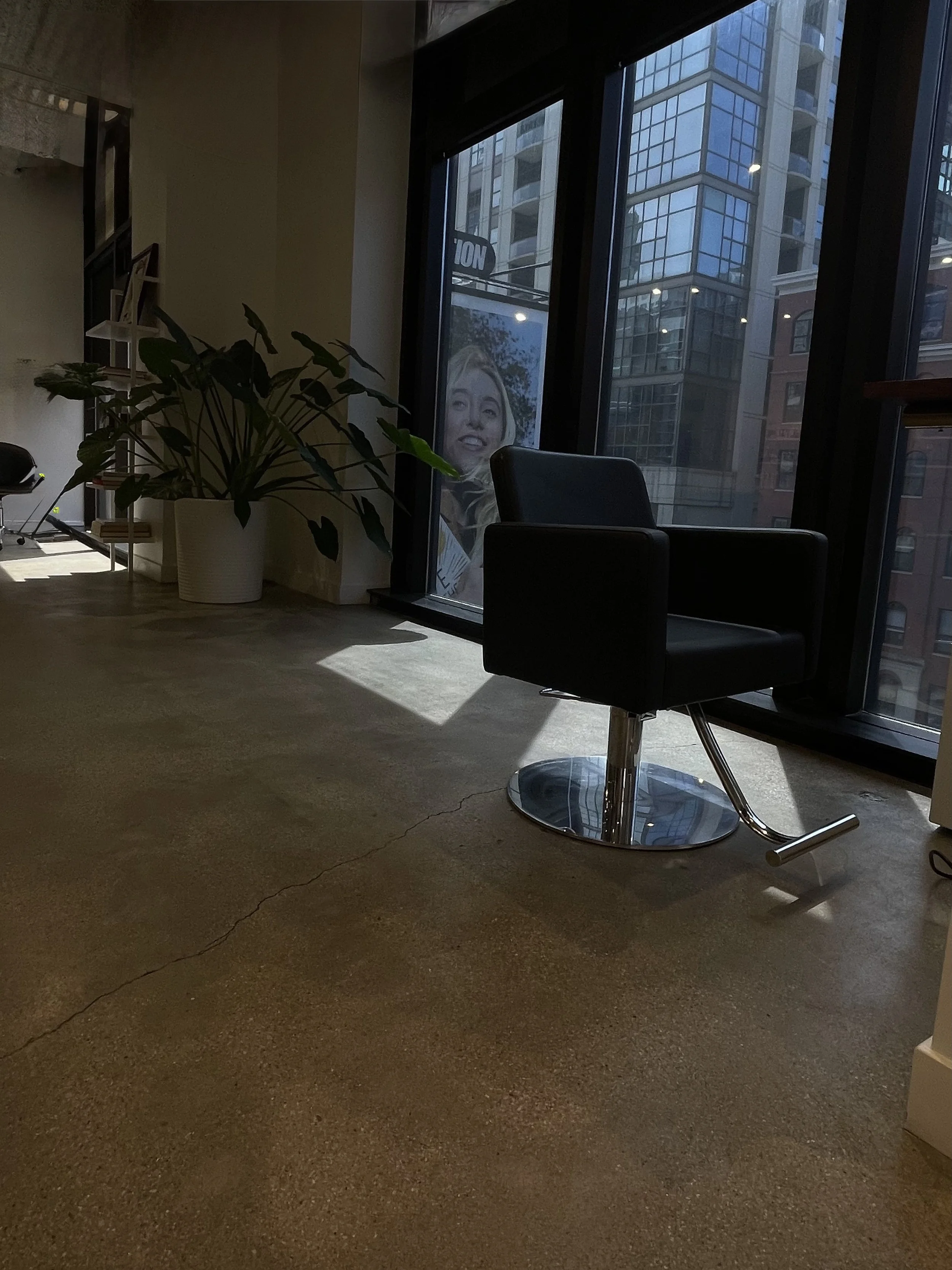 Interior of an office space with a black chair, large windows showing tall buildings outside, a potted plant, and sunlight casting shadows on the floor.
