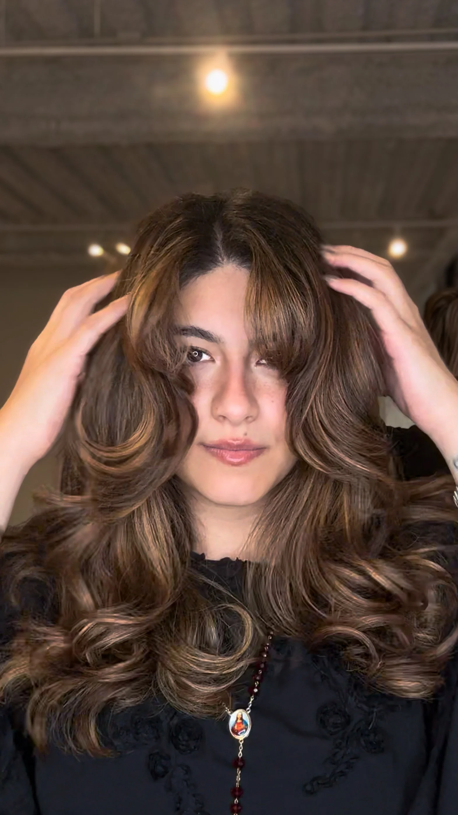 Woman with long, voluminous, curly hair, touching her hair, wearing a necklace with a religious pendant, and a black top.