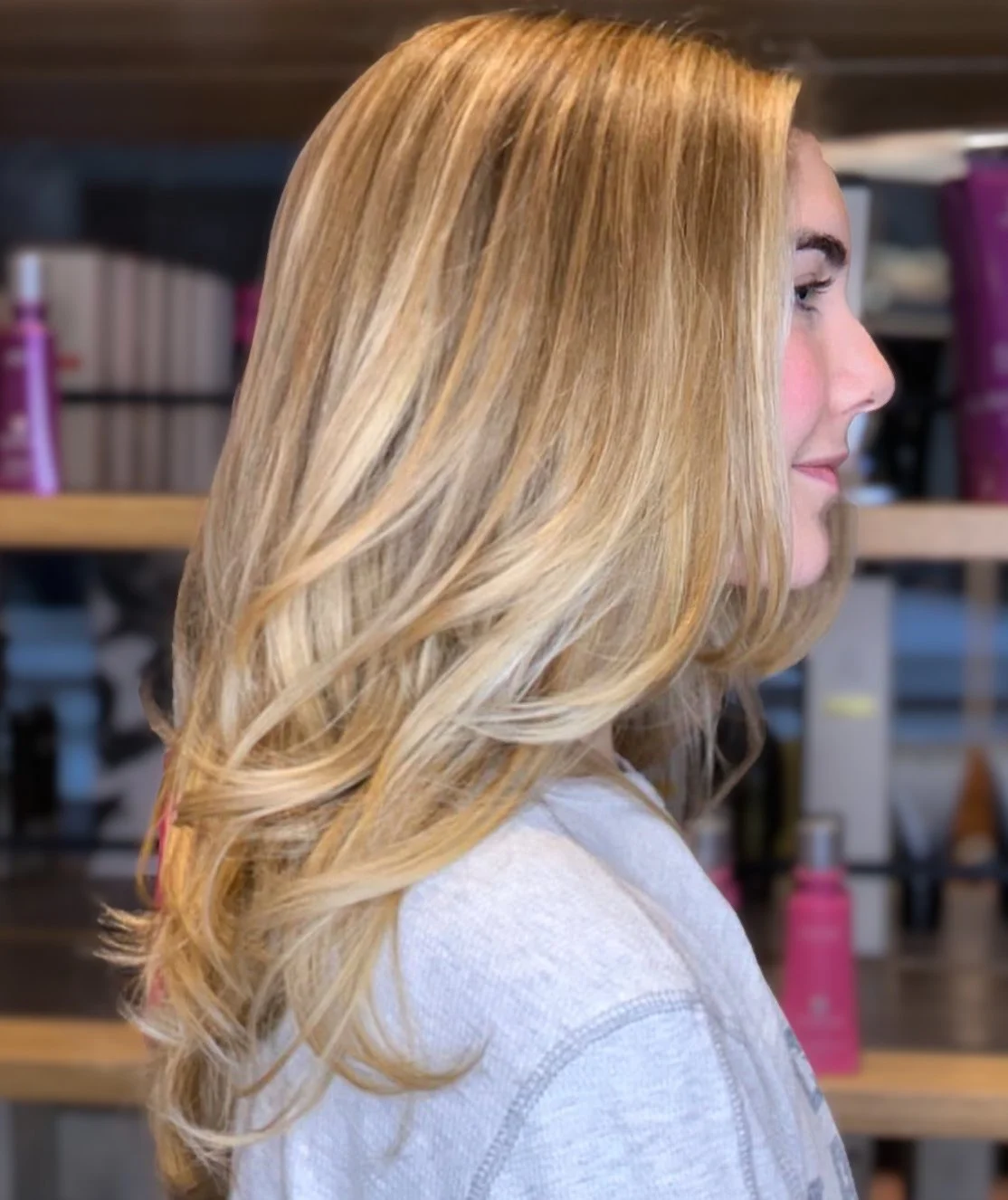 A woman with long, wavy blonde hair standing in a salon, with shelves of hair products in the background.