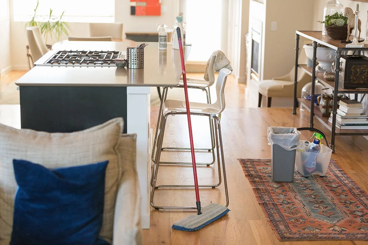 Kitchen with cleaning supplies, a mop, and a trash bin, adjacent to a sitting area with pillows and a shelving unit.