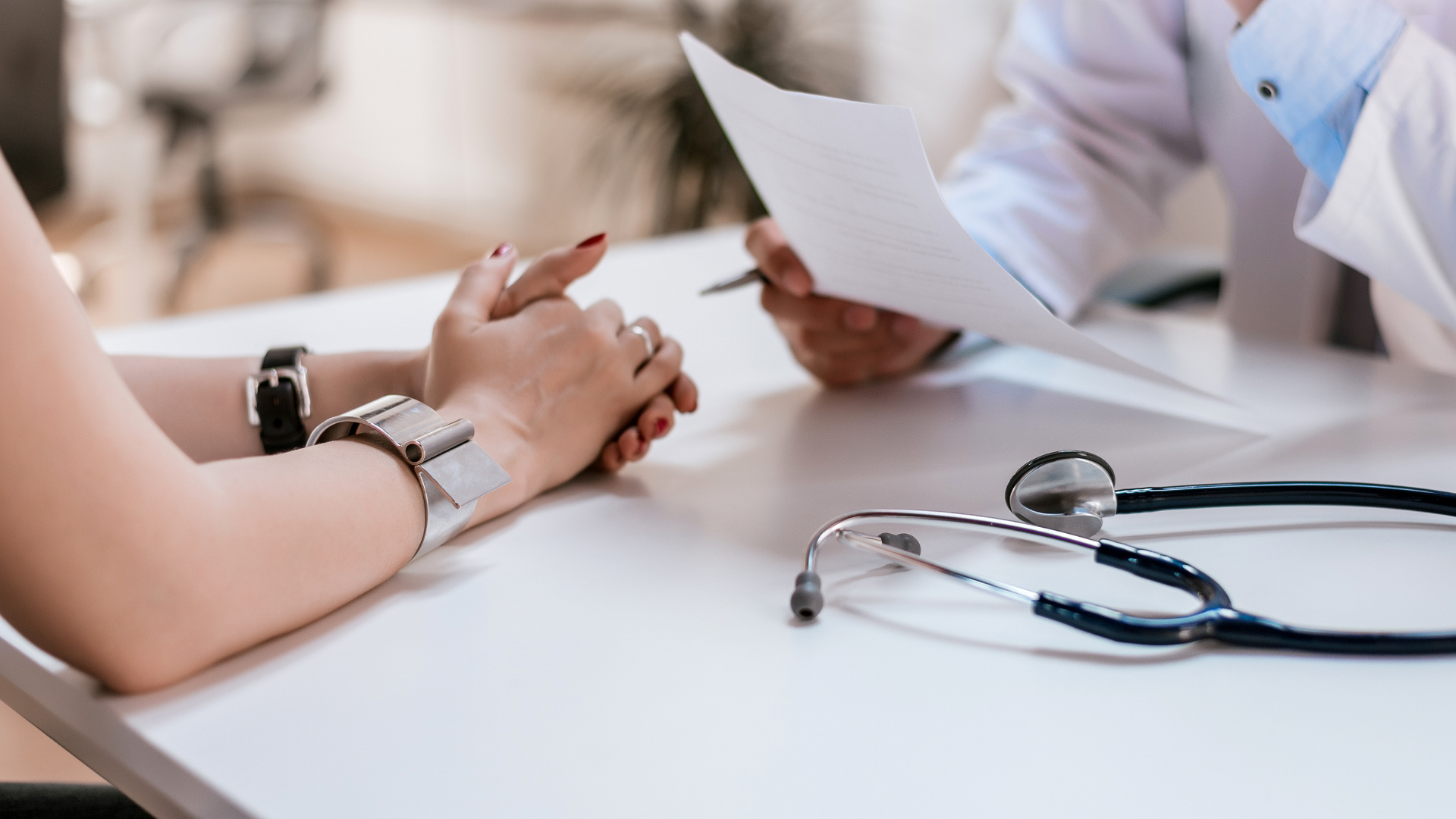 A doctor holding a clipboard and writing, while a patient with a watch and a bracelet holds their hands on a white table, with a stethoscope lying nearby.