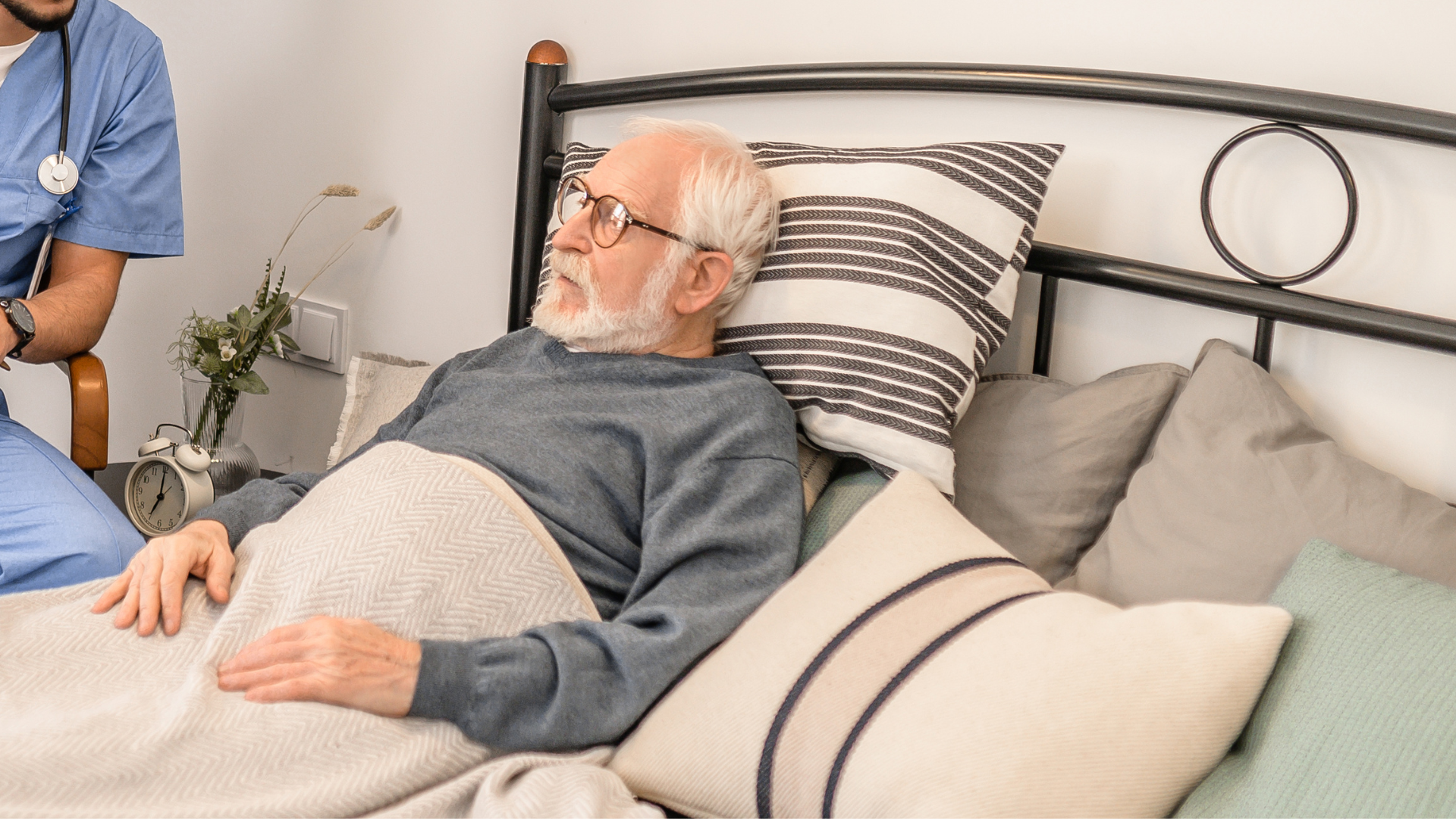 An elderly man with white hair, beard, glasses, wearing a gray sweater, is lying in bed with a blanket covering his lower body. A nurse in blue scrubs is sitting nearby, with only part of her arm visible. The bed is against a white wall, with a pillow behind the man's head and additional pillows on the bed, including a striped pillow and a green pillow. There is a small bedside table with a vase of flowers and an alarm clock.