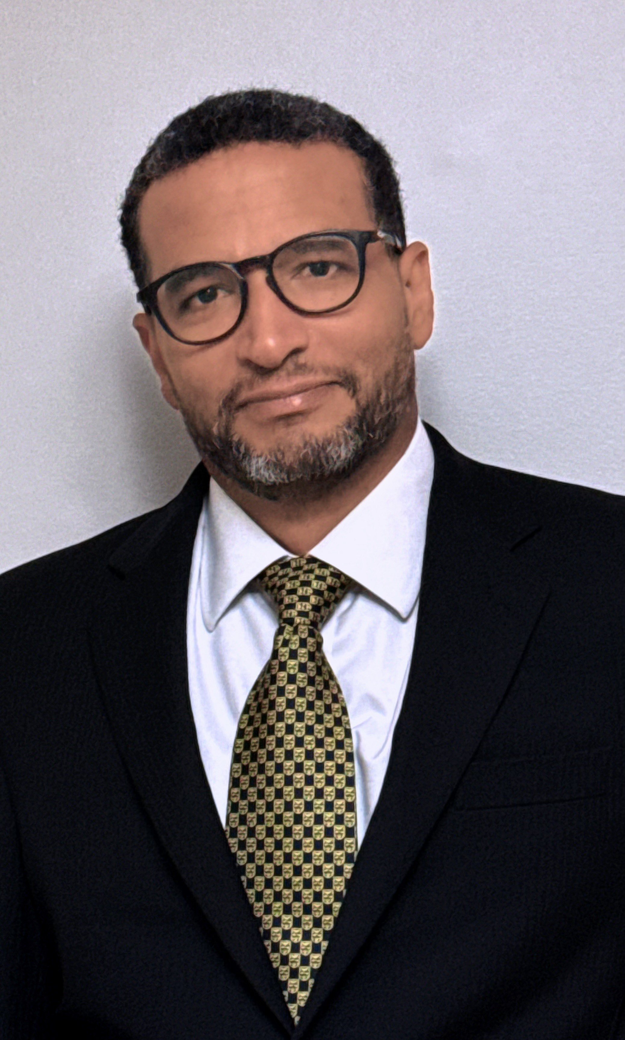 Man in a suit and tie, wearing glasses, posing against a plain background.