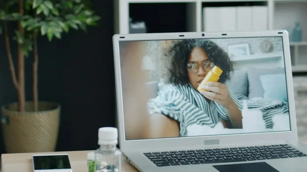 Laptop screen showing a woman with curly hair and glasses holding a bottle of medicine or lotion.