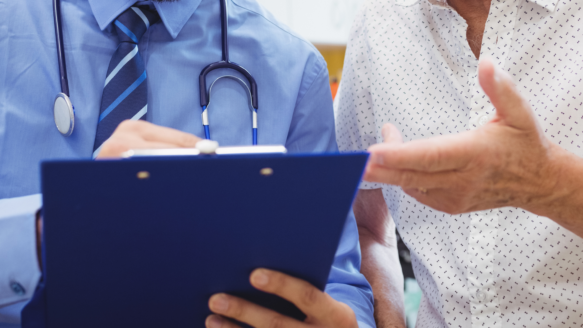 A doctor with a stethoscope around his neck talking to a patient, holding a clipboard, indoors.