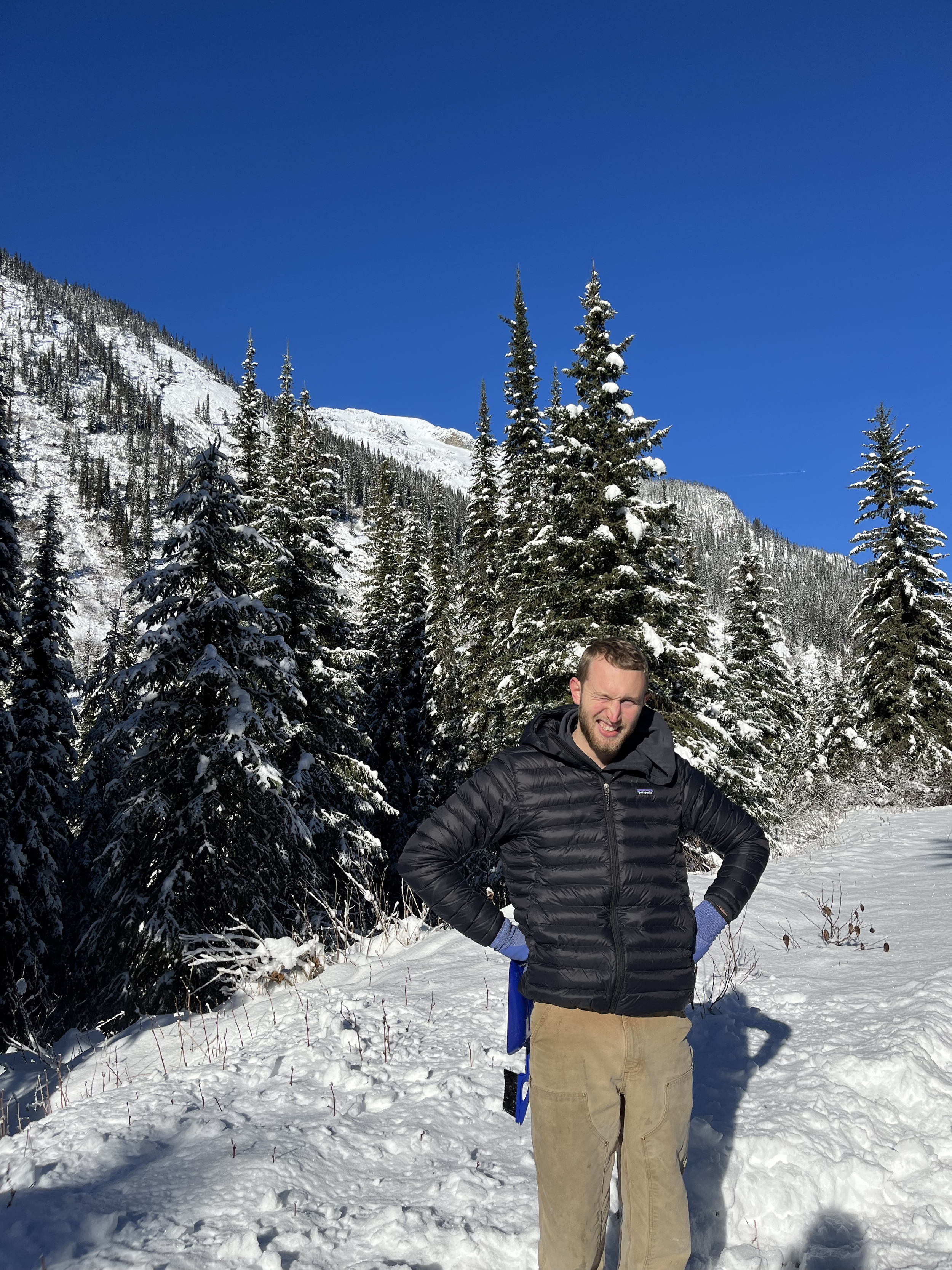 A man standing outdoors in a snowy forest with pine trees and mountains in the background, wearing a black jacket and beige pants.