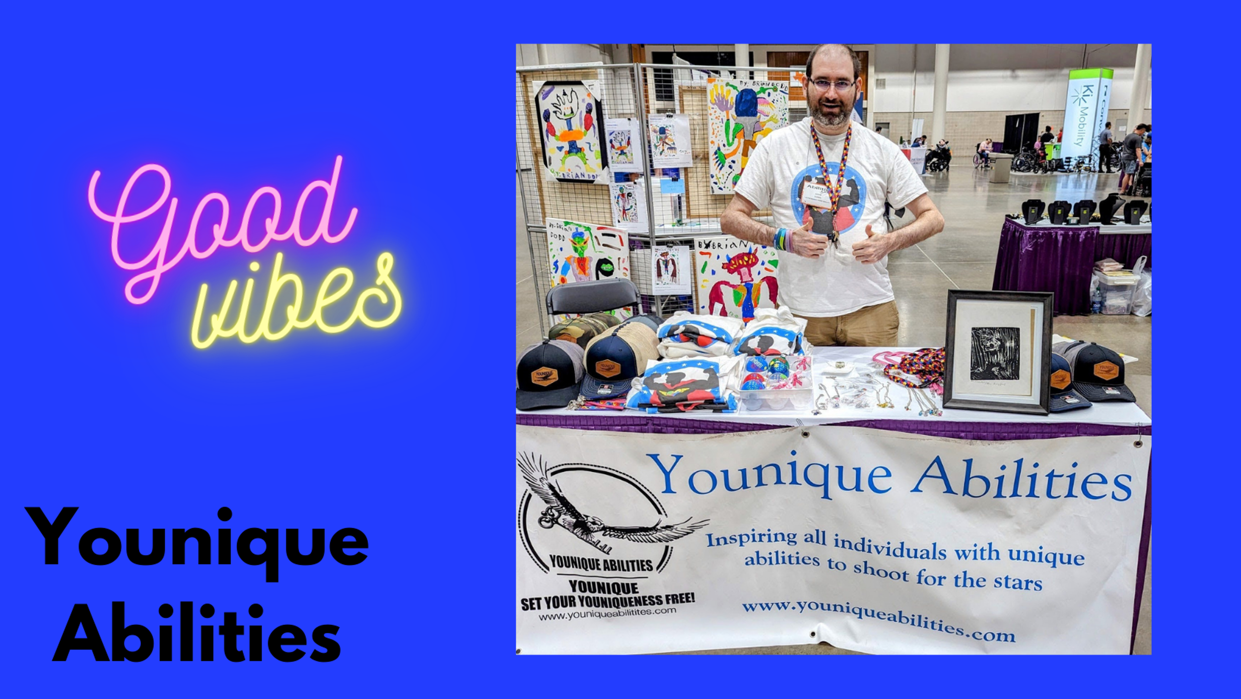 A man standing behind a table at a convention booth for Younique Abilities, displaying hats, T-shirts, jewelry, and artwork. The booth has a banner with the company's logo and website, with colorful artwork in the background and a neon sign that says 'Good Vibes'.