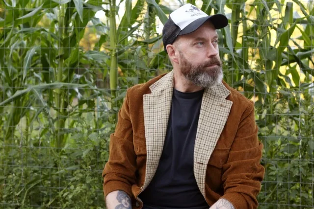 A man with a beard, wearing a baseball cap and a brown jacket, sitting outdoors in front of a fence and tall green plants.